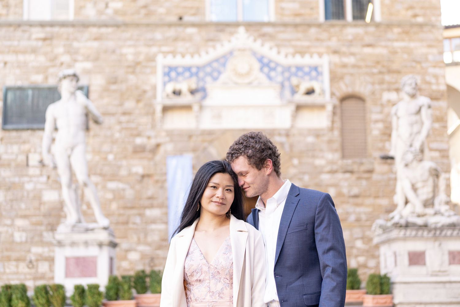 A couple in formal attire stands close together in front of an ornate stone building with statues.