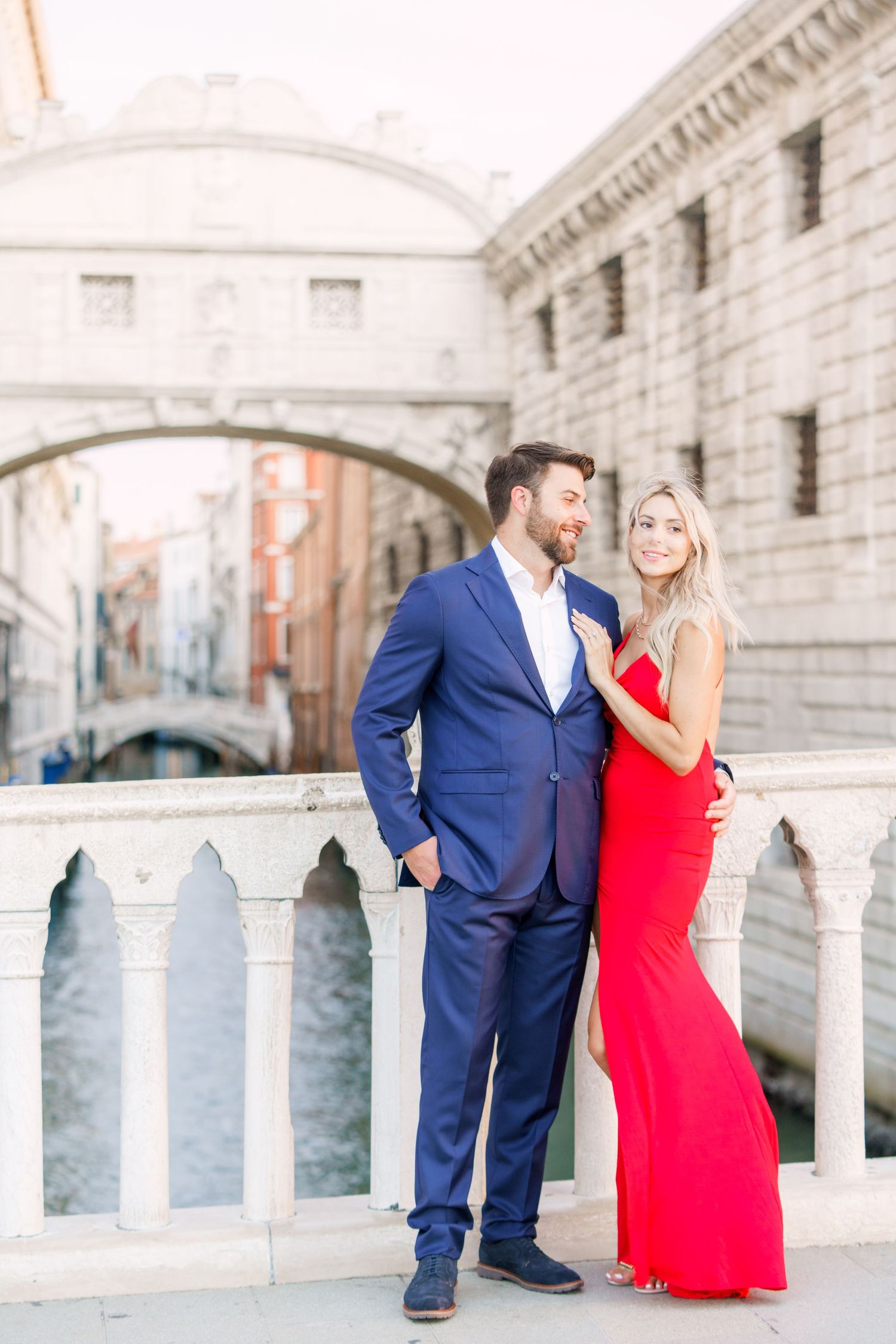 A couple poses together at the Bridge of Sighs in Venice, with the woman wearing a vibrant red dress and man in navy suit.