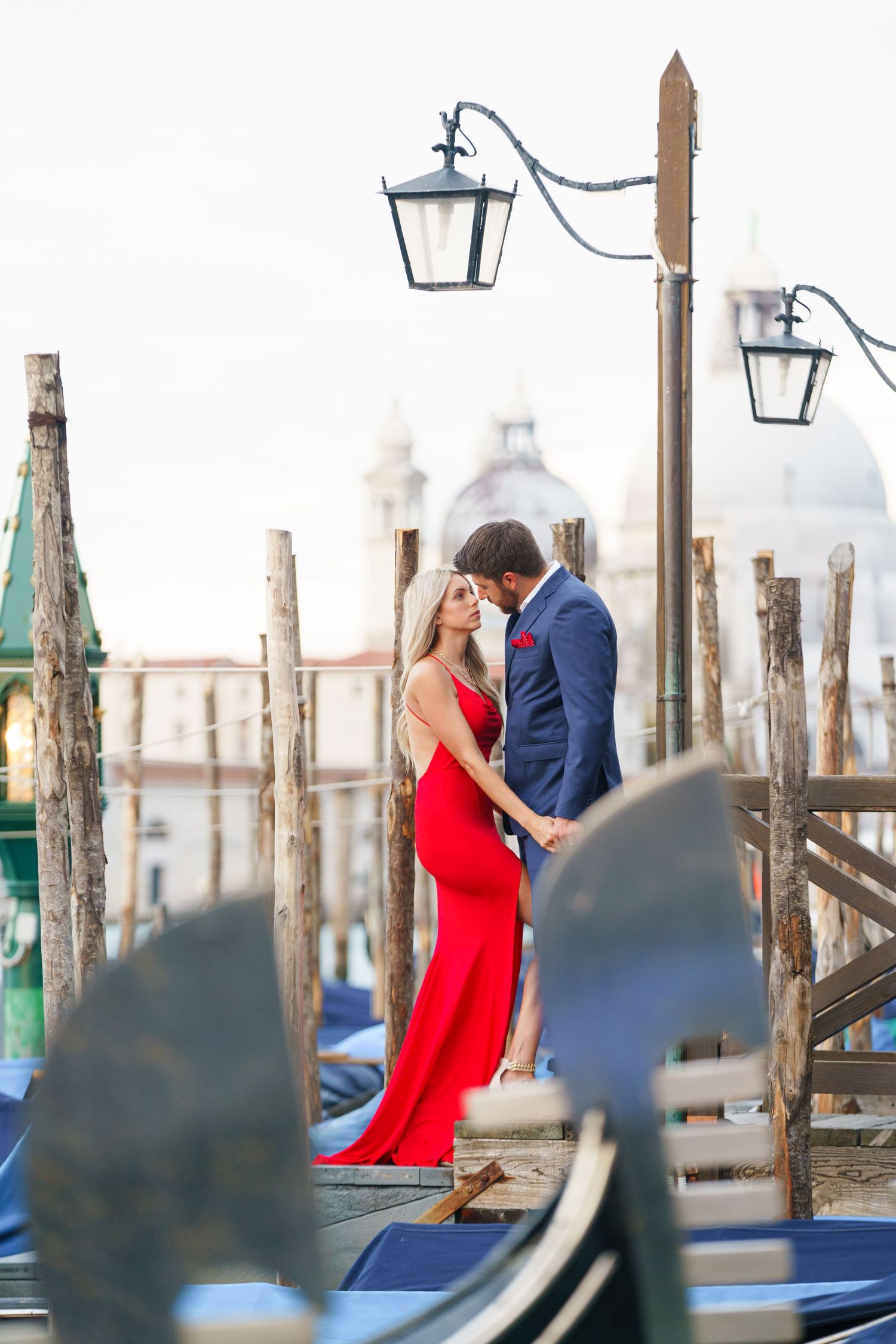 Romantic couple embraces on Venice dock with gondolas and lanterns at sunset, iconic Italian architecture in background.