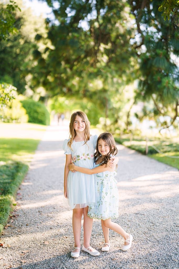 Two young girls in light blue dresses stand together on a gravel path surrounded by greenery on a sunny day.