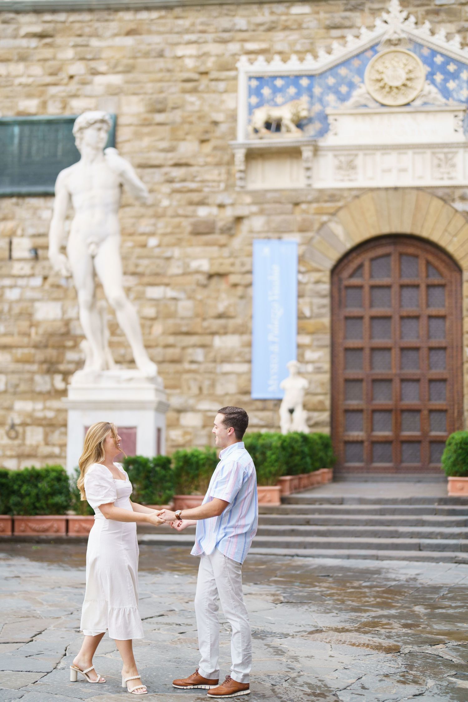A couple holds hands in front of a historic stone building with a classical statue and ornate architectural details in Florence.