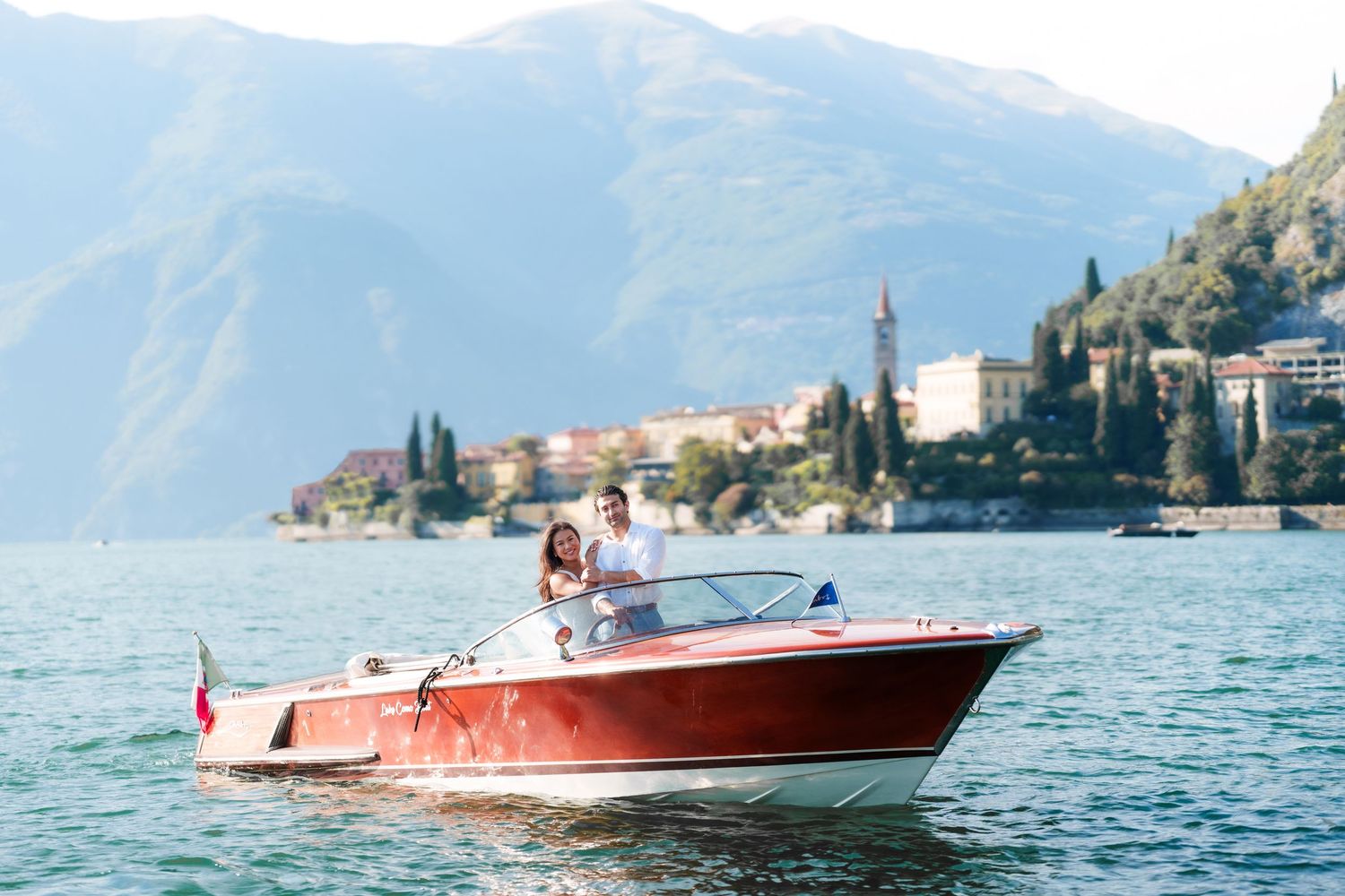 Couple riding a classic red wooden boat on Lake Como with mountain village backdrop on a sunny day.