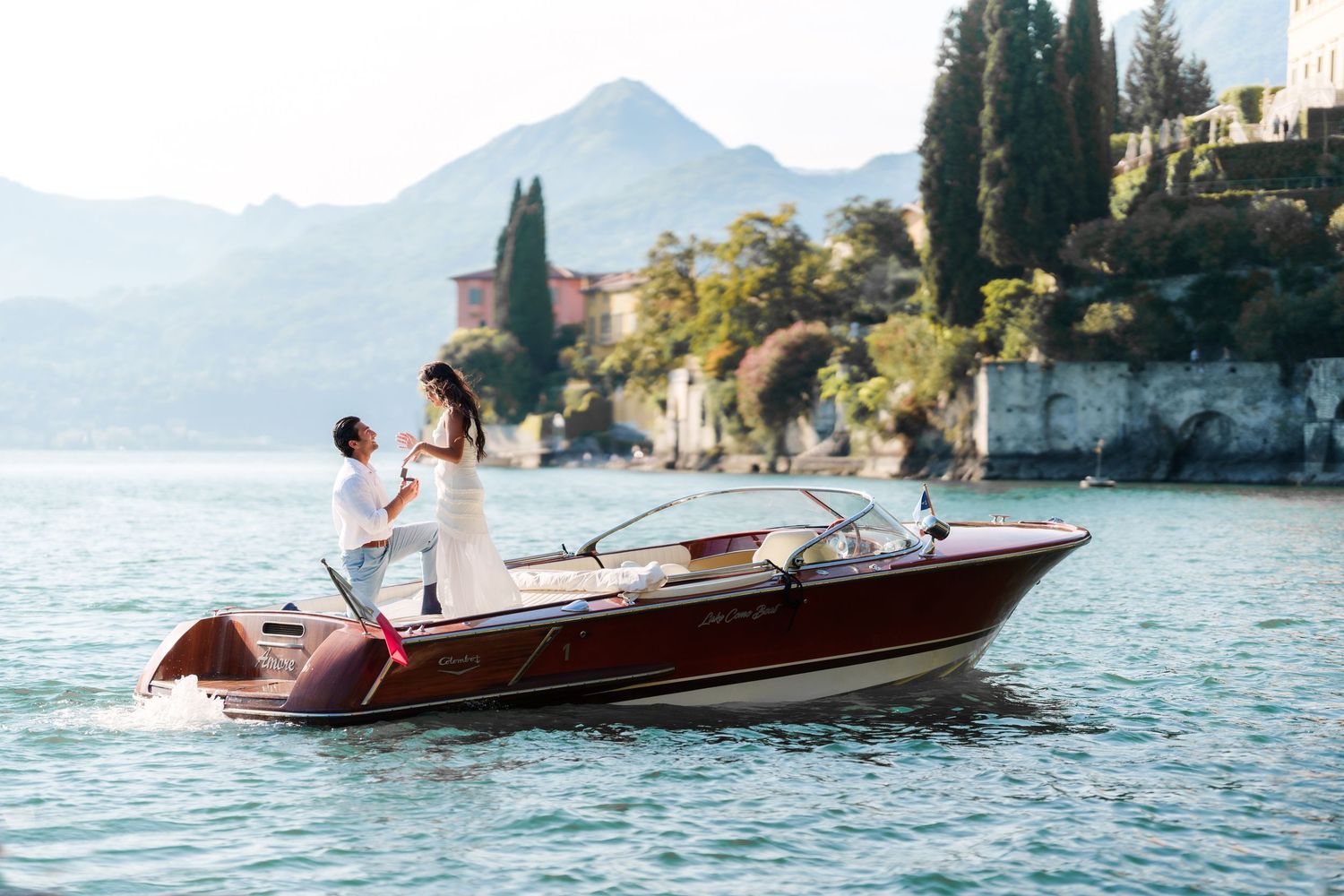 Couple in wedding attire standing on a classic wooden speedboat on a serene lake with mountains and villa backdrop.