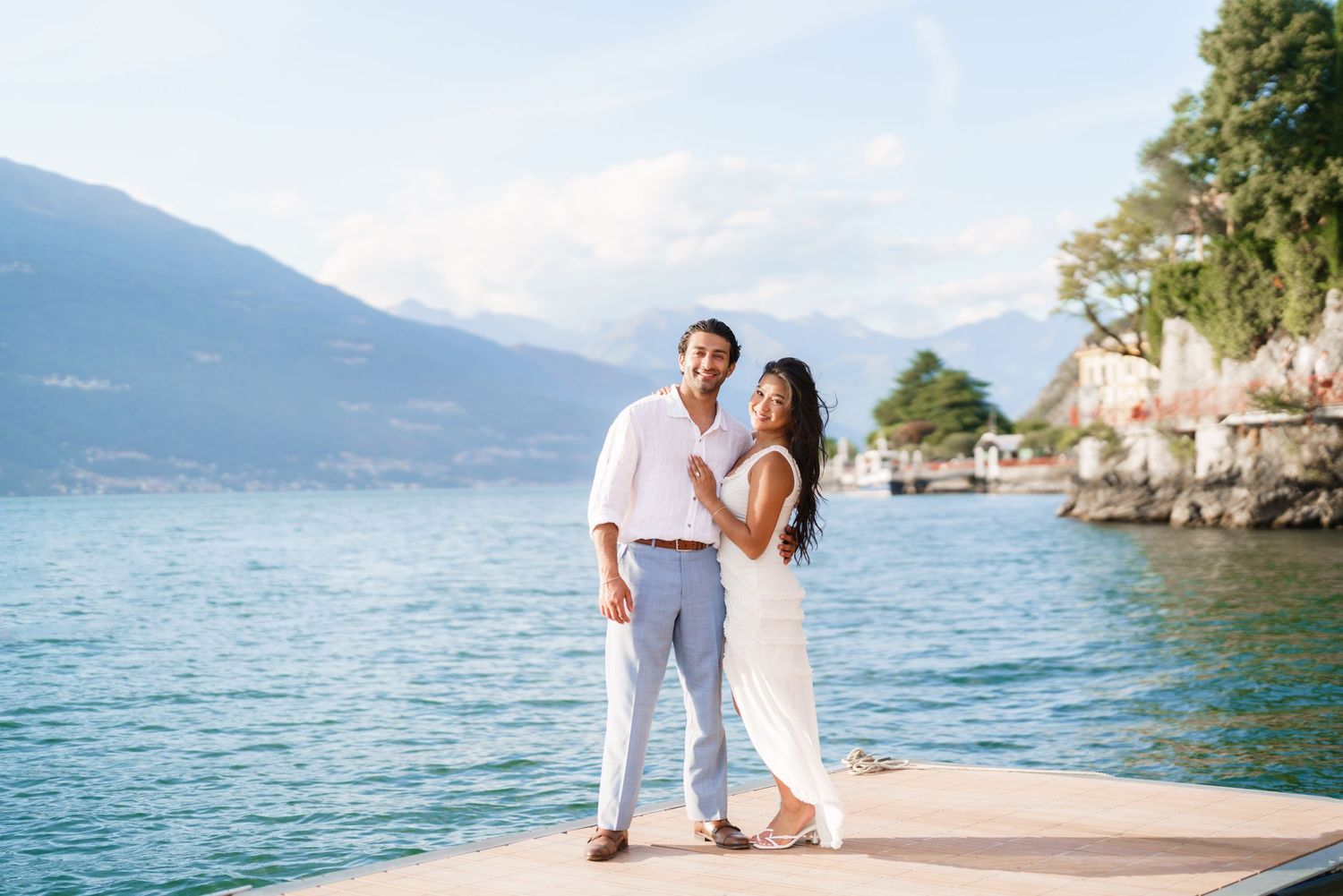 Couple in white outfits posing on a dock by a serene lake with mountains and lush greenery in the background.