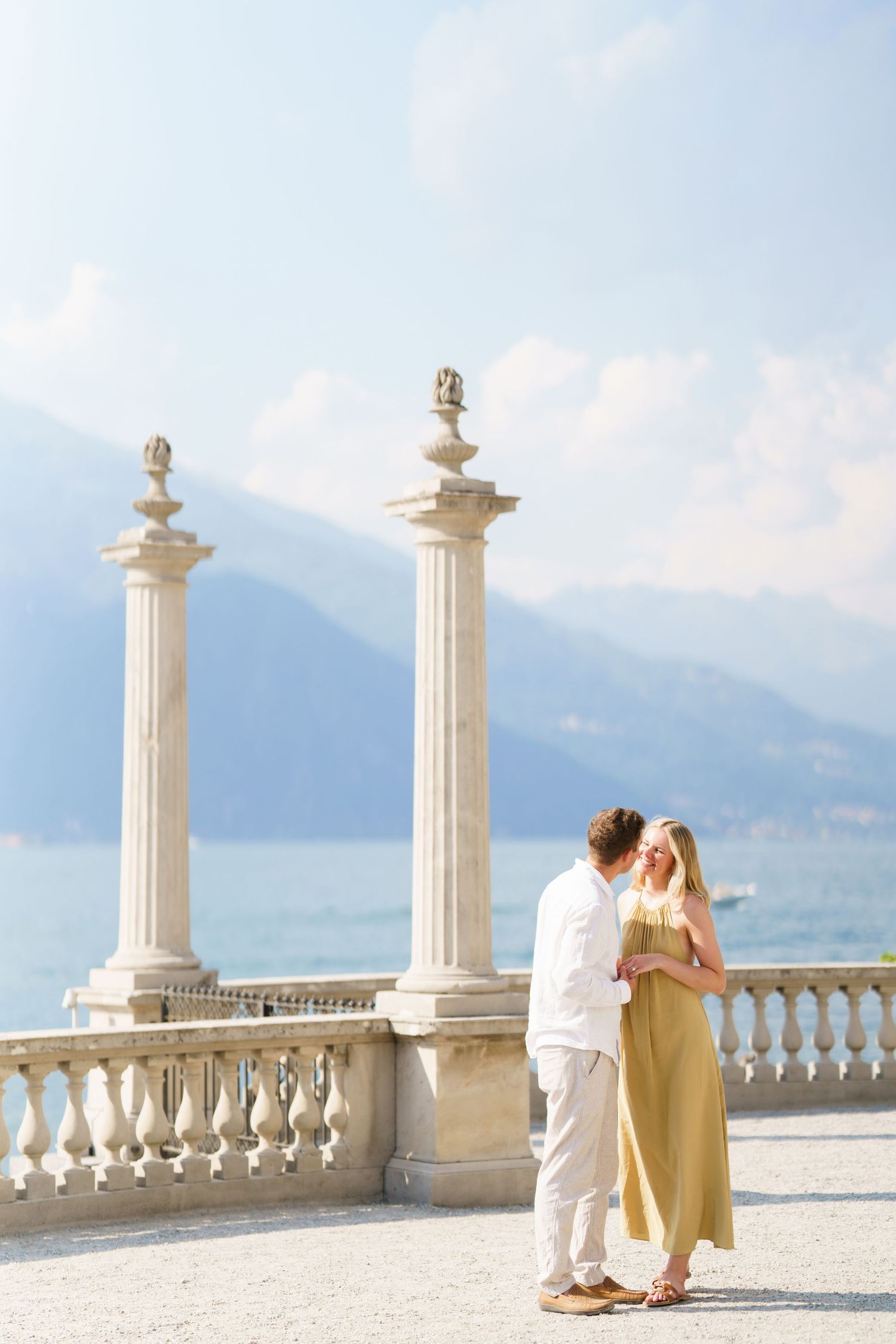Couple embracing on elegant lakeside terrace with classical columns and mountain backdrop.