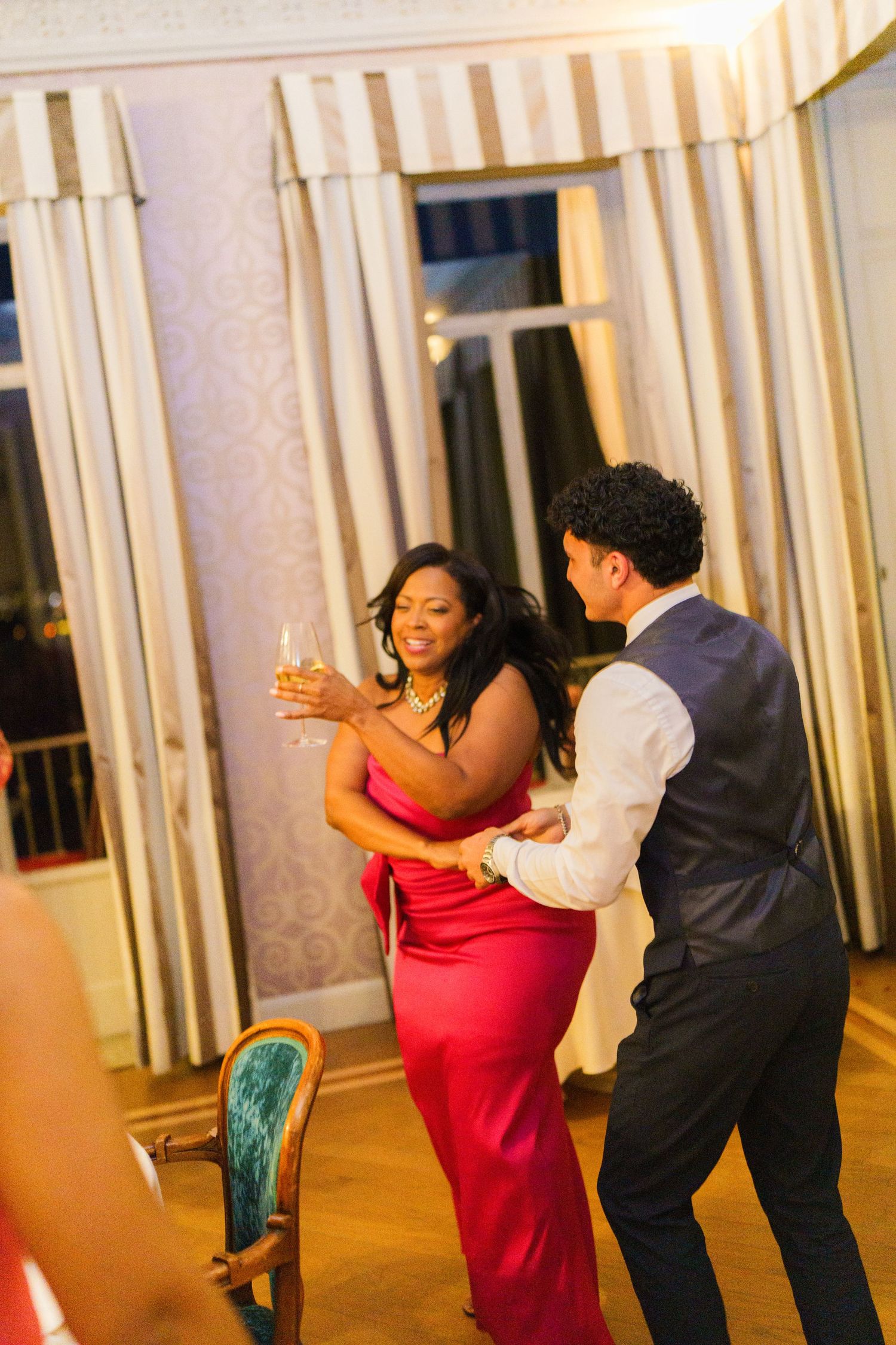 Couple dancing at elegant reception, woman in red gown holding champagne, man in navy suit, warm lighting.