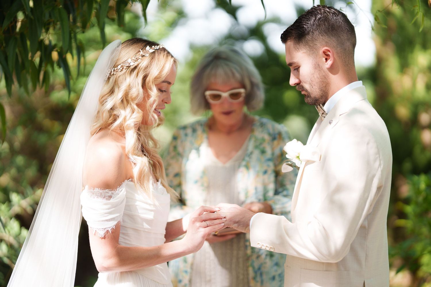 Bride placing wedding ring on groom's finger during outdoor ceremony with officiant in floral dress.