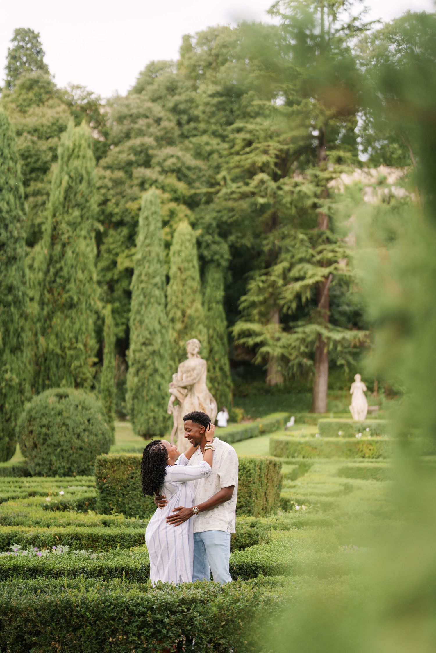 A couple embraces in a formal Italian garden with manicured hedges, cypress trees, and classical statues in the background.