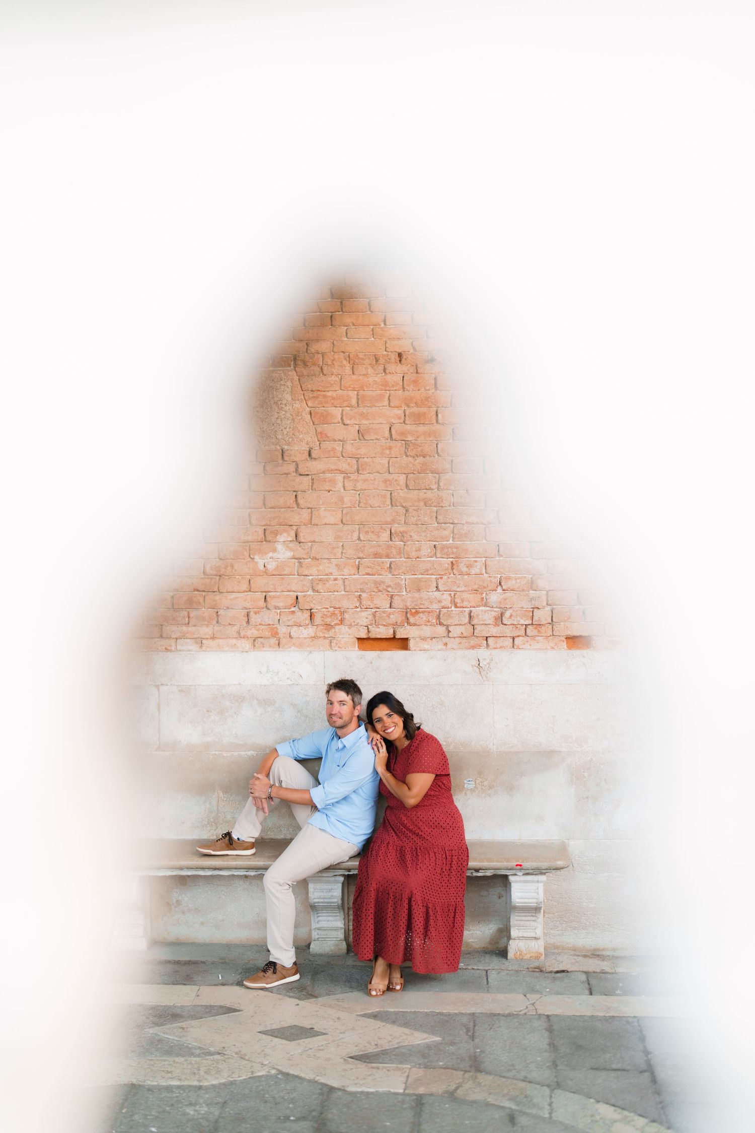 Couple posing romantically on a stone bench framed by a decorative archway with exposed brick wall backdrop.