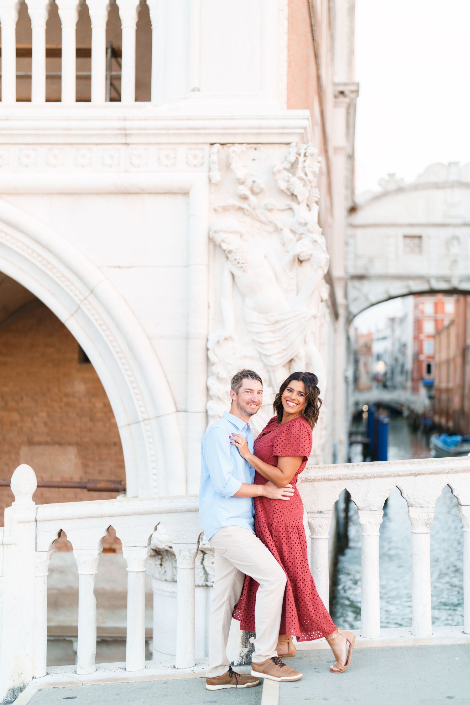 Couple embracing on a white marble bridge in Venice, Italy, with a canal and the Bridge of Sighs in the background.