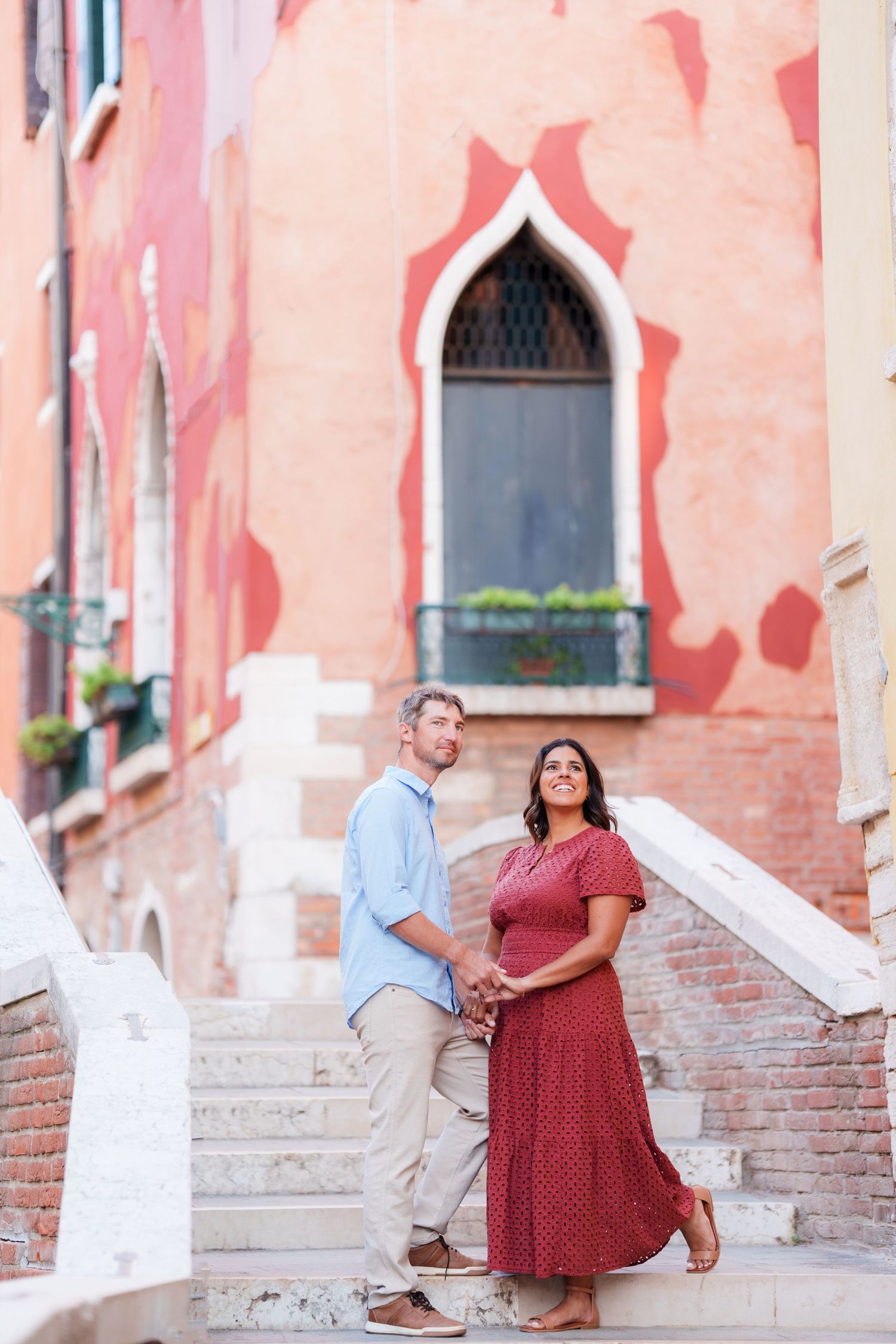 Couple holding hands on stone steps in Venice, Italy, with colorful red building facade in background.