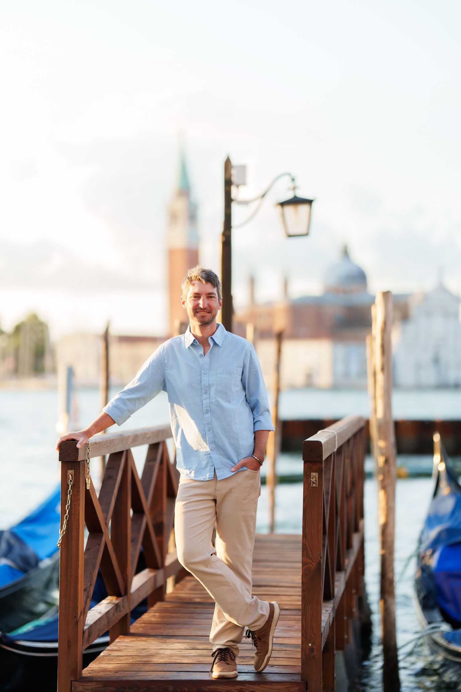 Man in blue shirt posing on wooden dock in Venice, Italy, with gondolas and San Giorgio Maggiore in background.