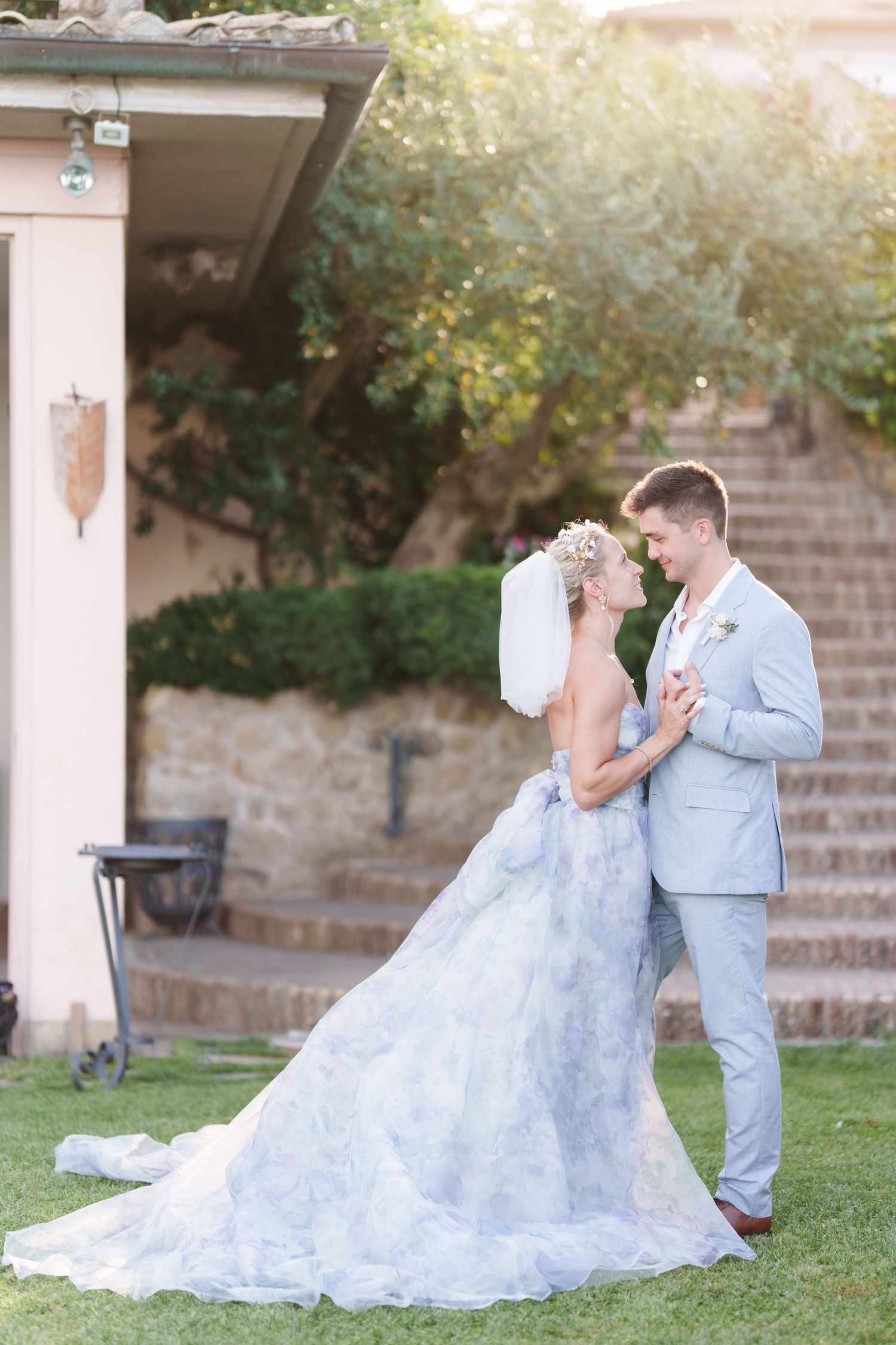 Bride in floral gown and groom in light blue suit share romantic moment on green lawn at elegant stone venue.
