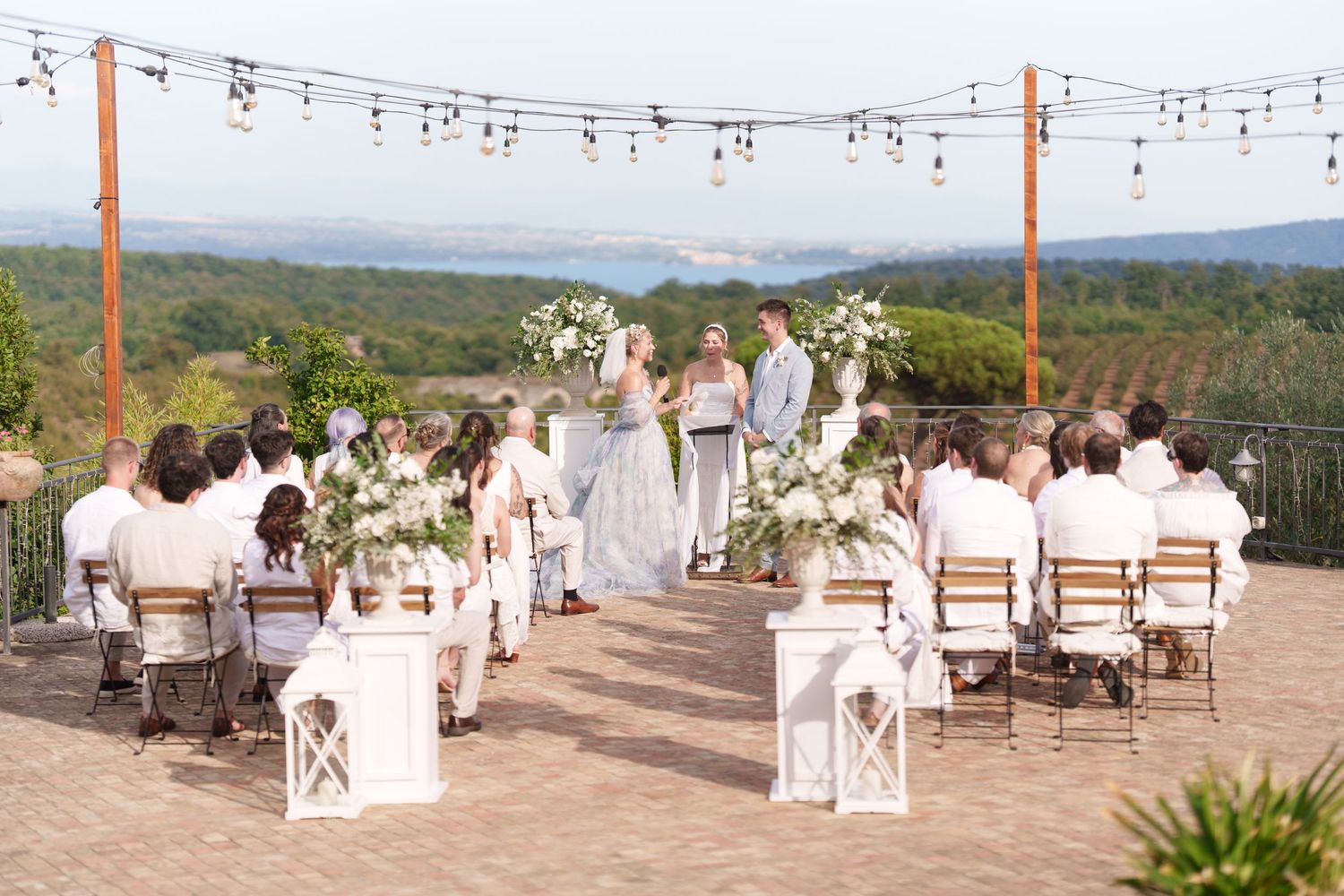 Outdoor wedding ceremony on a terrace with string lights, floral arch, and scenic valley views.