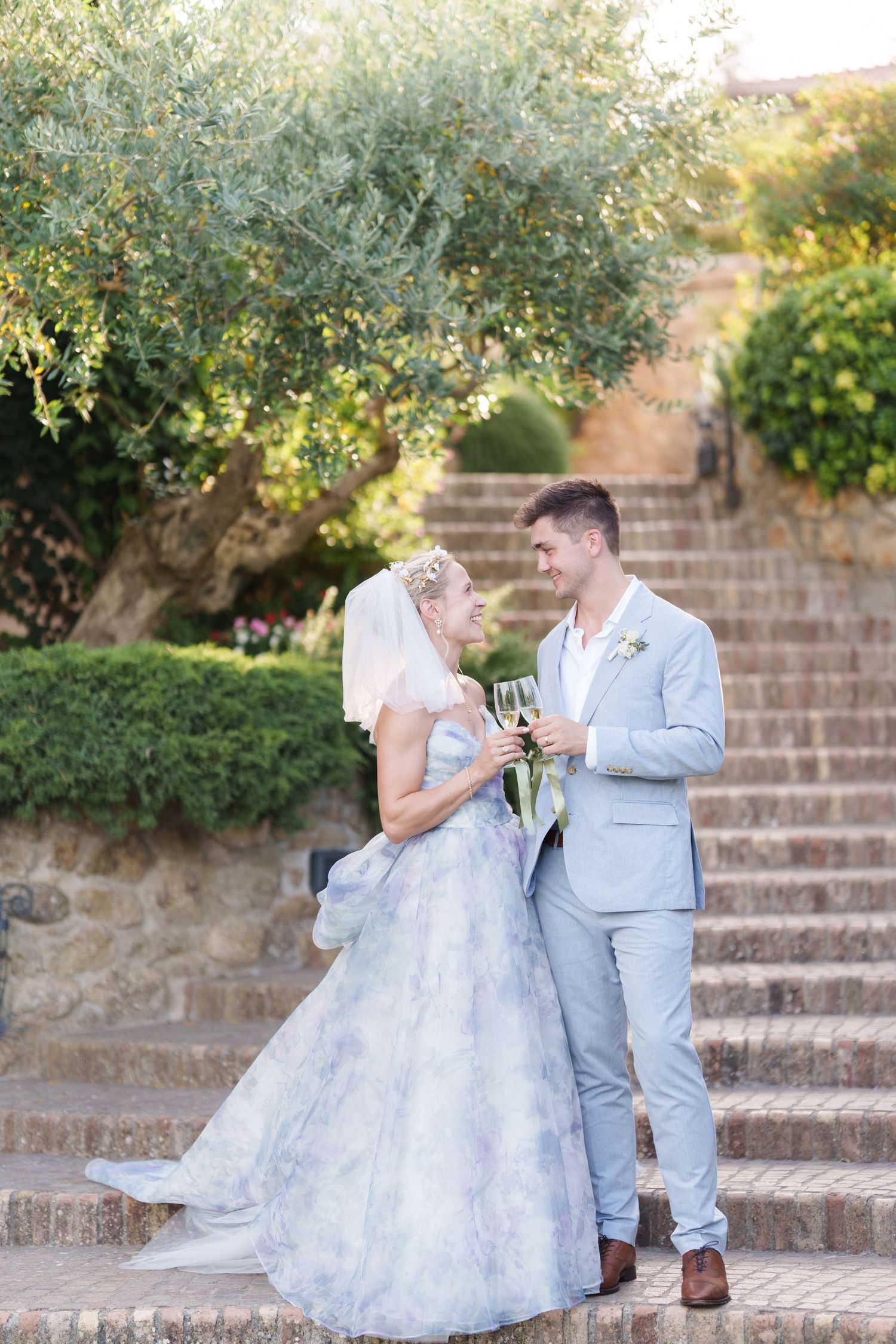 Bride in blue floral gown and groom in gray suit toasting champagne on stone steps surrounded by lush greenery.