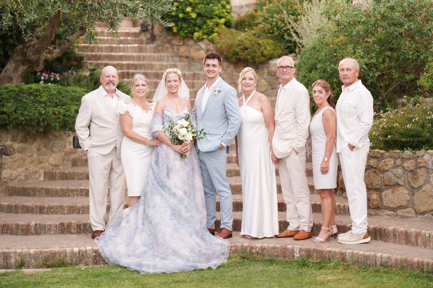 Wedding party poses on stone steps outdoors, bride in floral gown with bouquet, groom in light blue suit, surrounded by family.