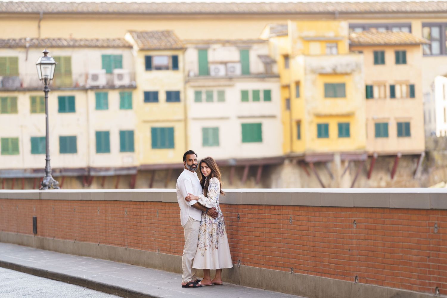 Couple embracing on a bridge in Florence, Italy, with colorful Ponte Vecchio buildings in the background.