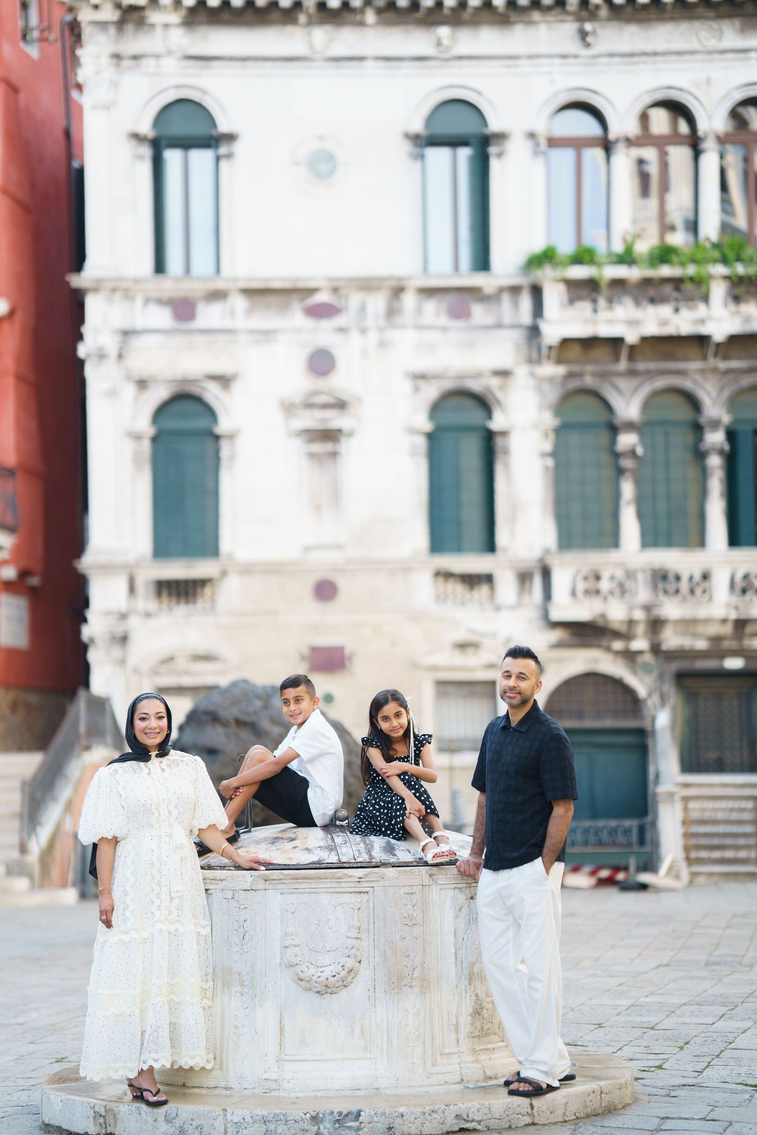 Family of four posing by a stone well in a Venetian piazza, with ornate historic buildings in the background.