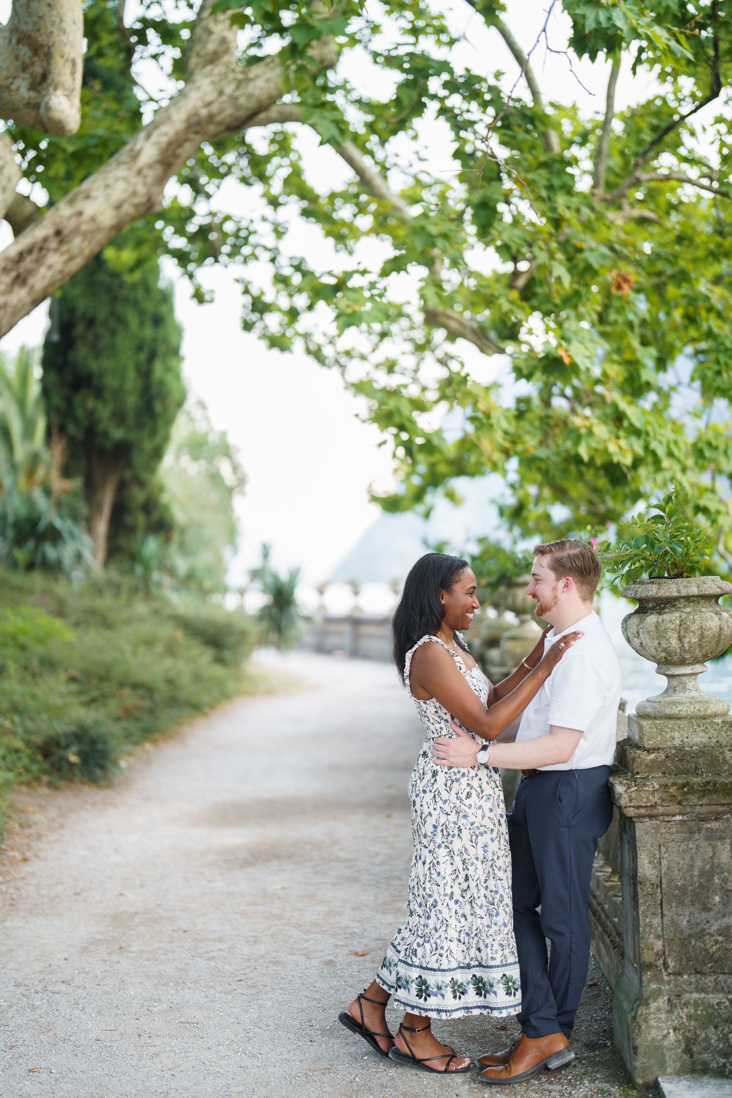 Romantic interracial couple gazing at each other by a stone balustrade on a garden pathway.