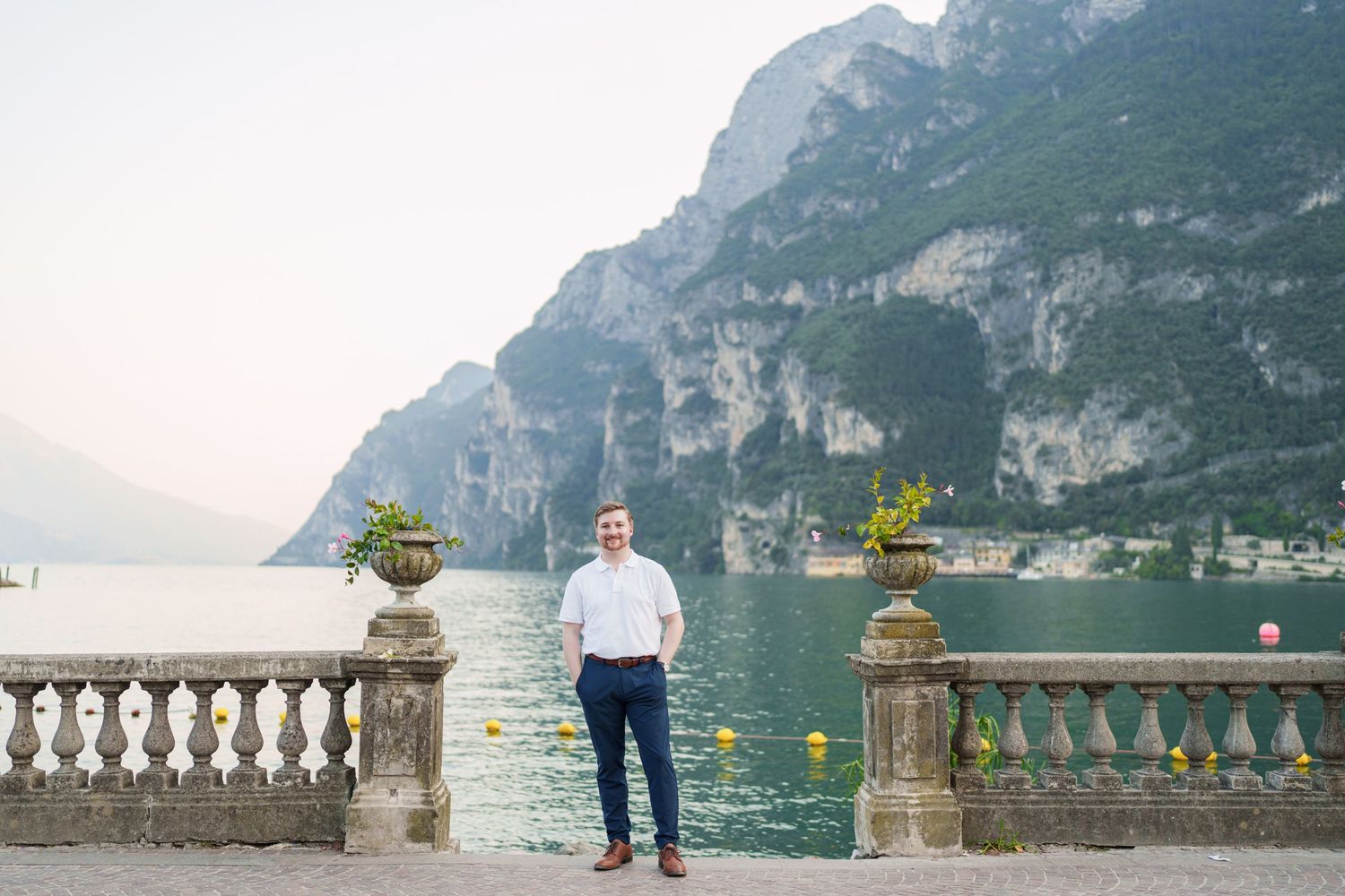 Smiling man standing on elegant stone balustrade terrace overlooking turquoise Lake Garda with mountain cliffs.
