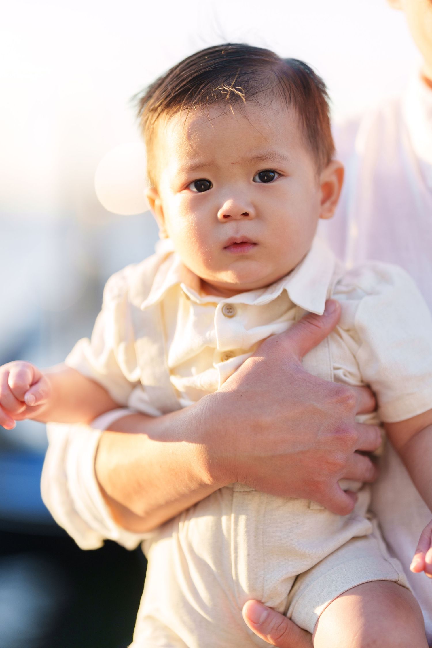 Adorable Asian baby boy in cream outfit being held by adult hands in warm sunlit outdoor setting.