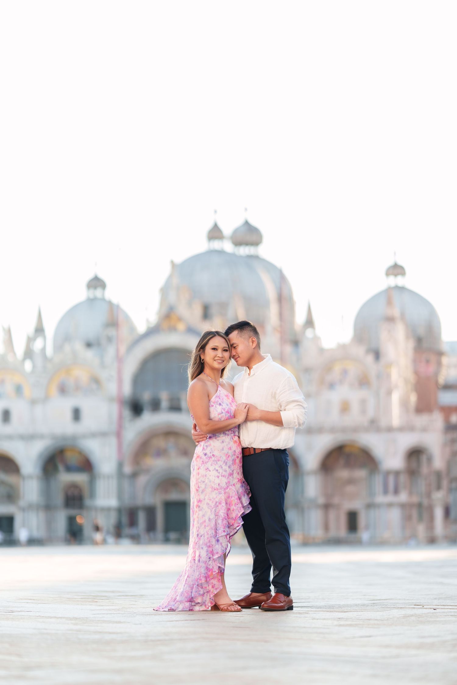 Couple embracing in St. Mark's Square, Venice, woman in pink floral dress, man in light shirt, Basilica backdrop.