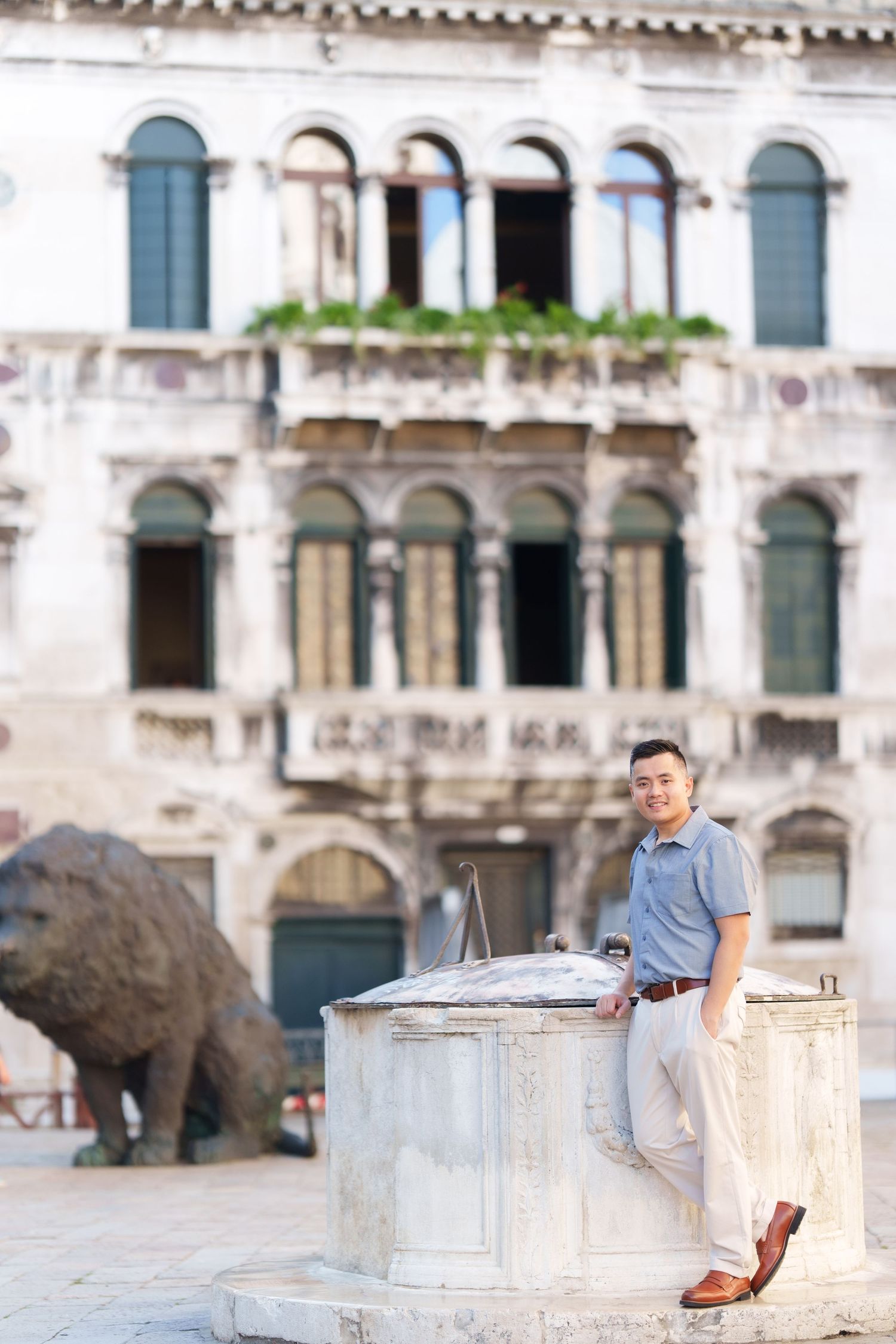 Man leaning against a stone well in a historic Venetian square with ornate building facade and lion statue behind him.