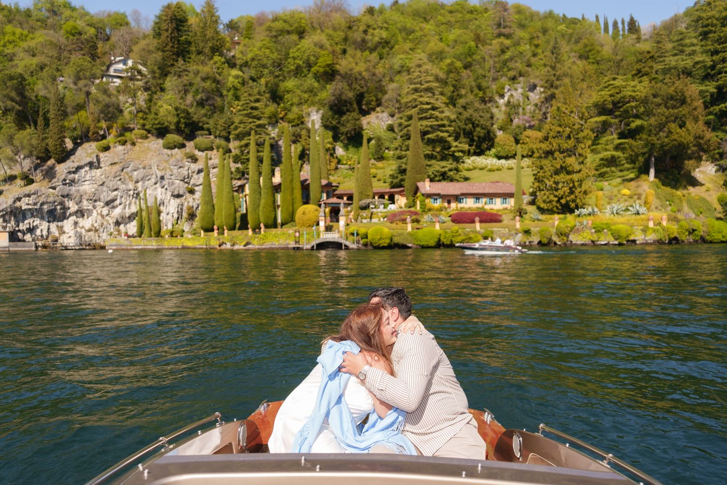 Couple kissing on a boat with lush green hillside villa and cypress trees on Lake Como in background.