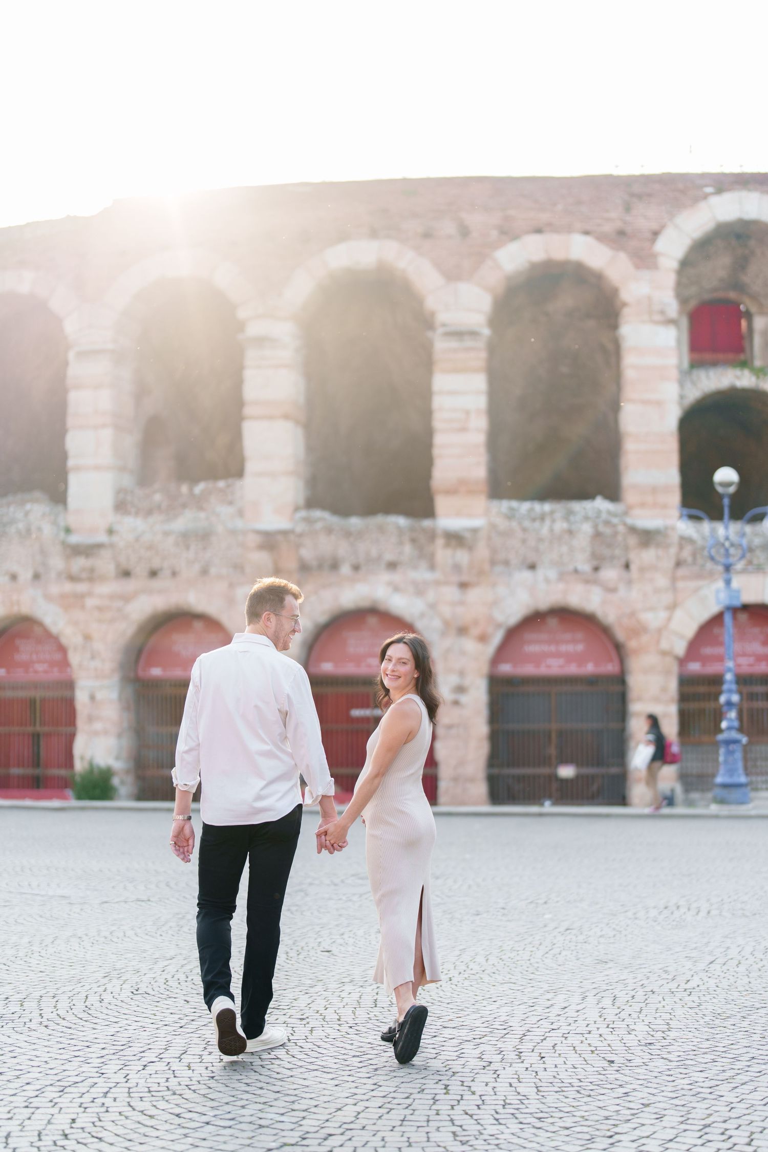 A couple walks hand in hand on cobblestone streets in front of the historic Verona Arena in Italy during sunset.