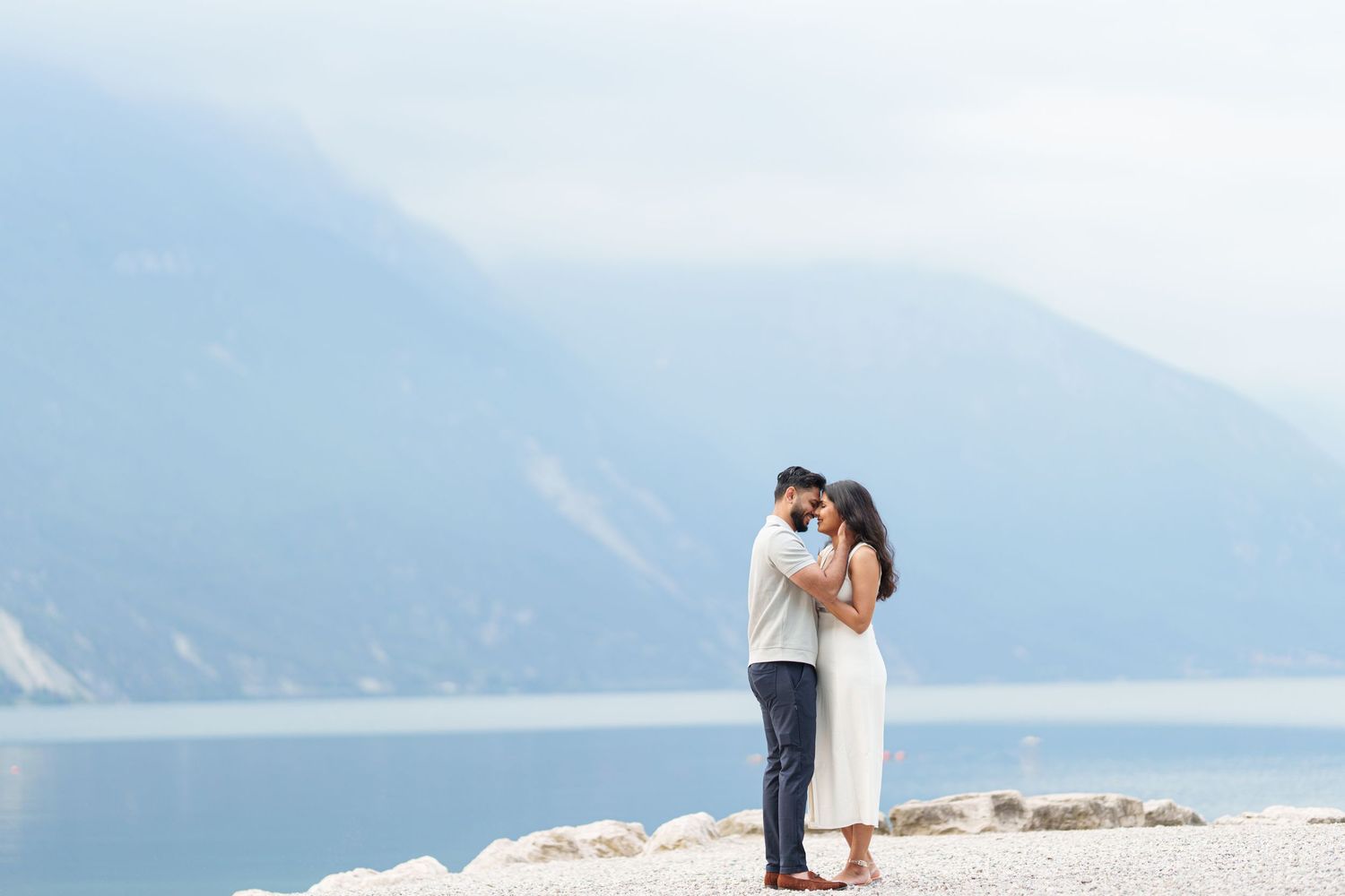 A couple embraces while standing on a mountain overlook with a scenic lake and mountain vista in the background.