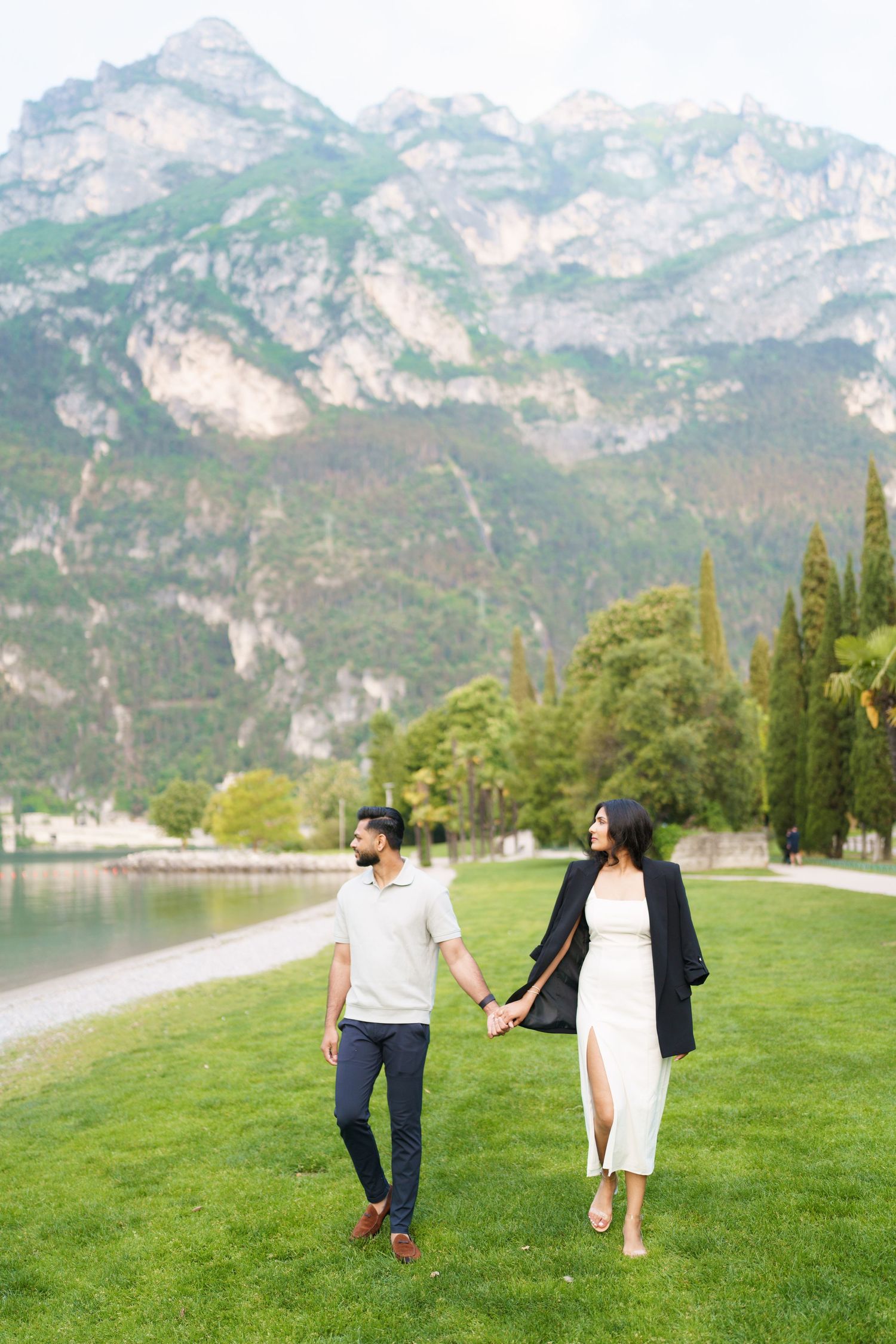 A couple walks hand in hand on a lush green hillside with majestic mountain peaks rising in the background.
