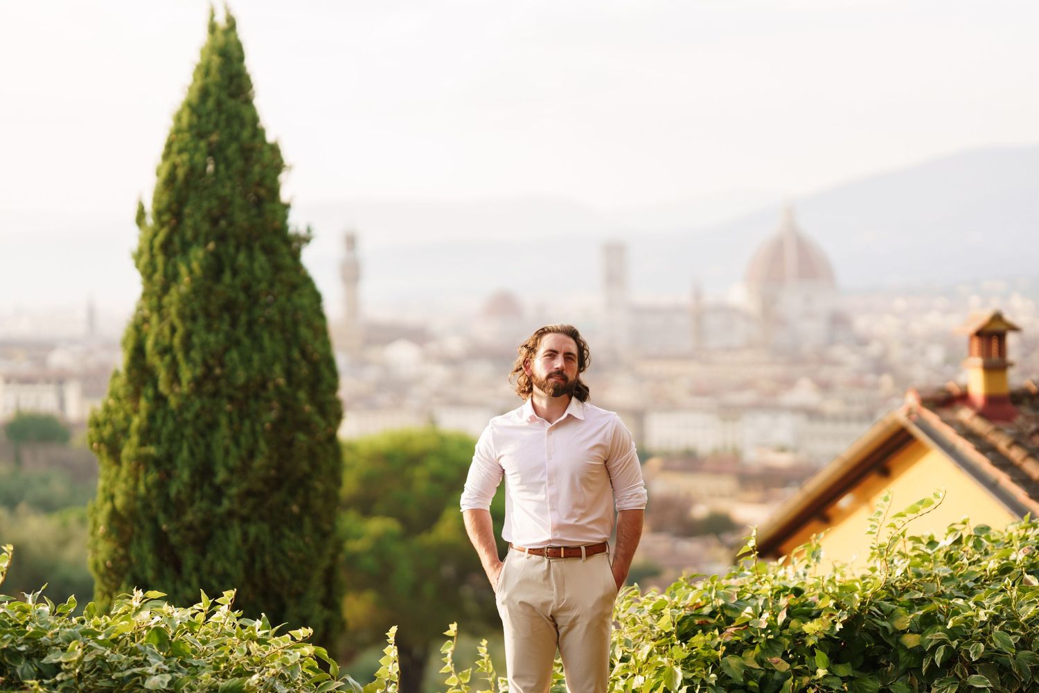 A scenic view of a Mediterranean hillside with cypress trees and buildings overlooking a distant cityscape.
