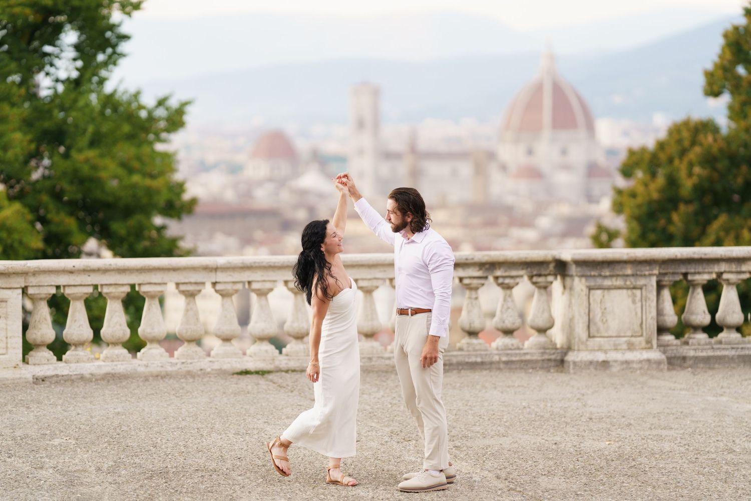 A couple in white outfits walks hand in hand along a scenic overlook with Florence's Duomo cathedral visible in the distance.