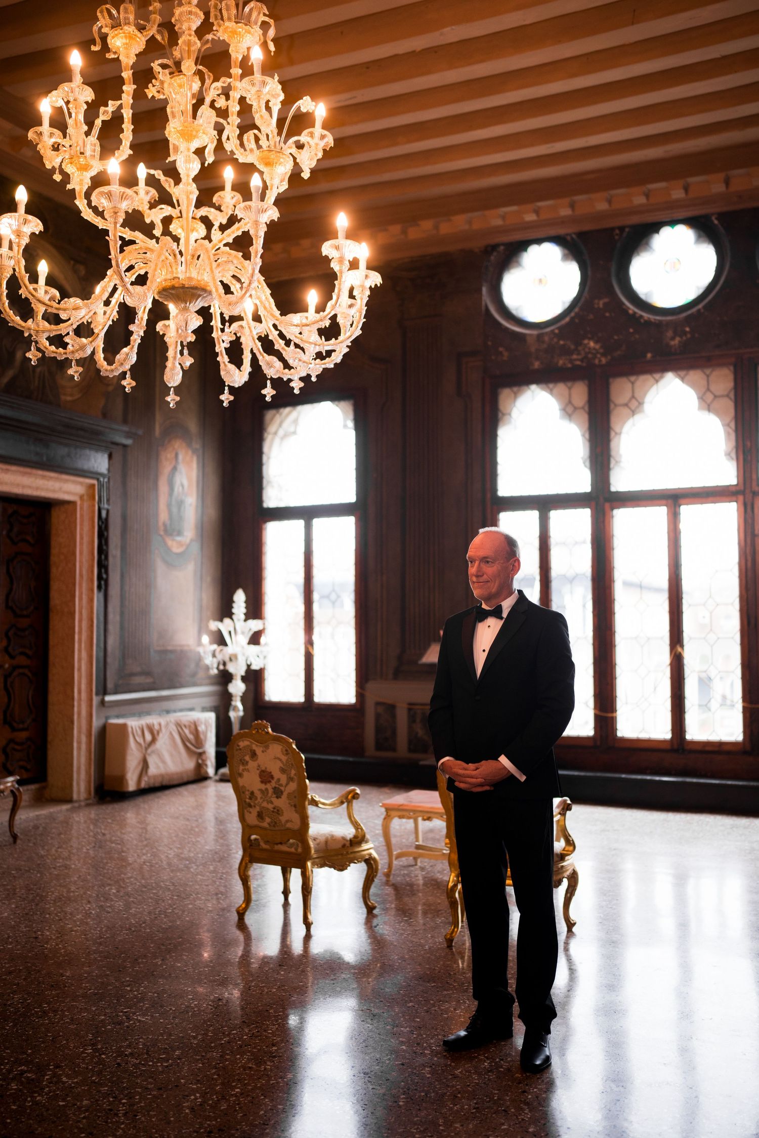 A silhouetted figure stands in an elegant room with ornate chandeliers and large windows casting dramatic lighting.