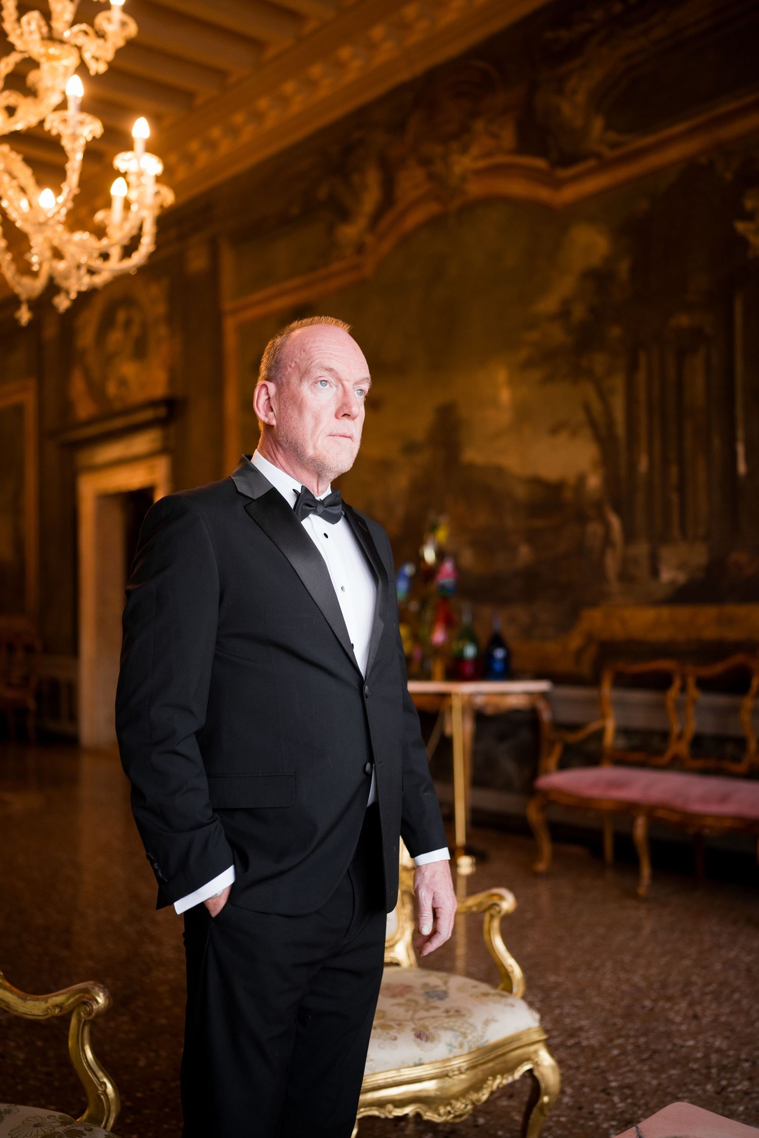 Man in black tuxedo standing in ornate room with chandeliers and gilded furniture.