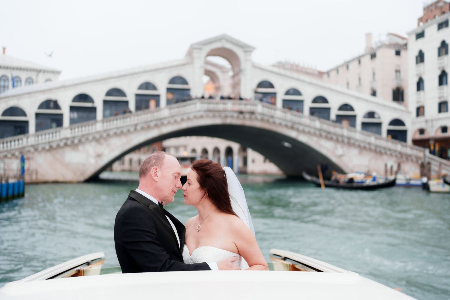 Romantic couple shares a kiss while riding in a gondola along Venice's Grand Canal with Rialto Bridge in background.