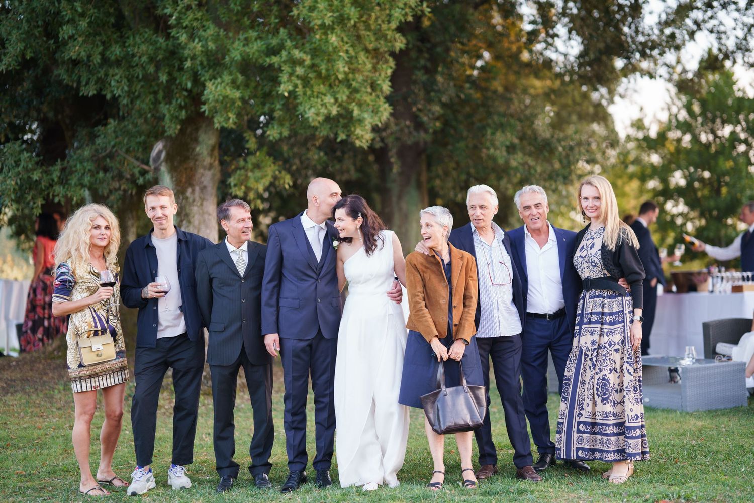 Large wedding party group poses together under trees at outdoor venue.