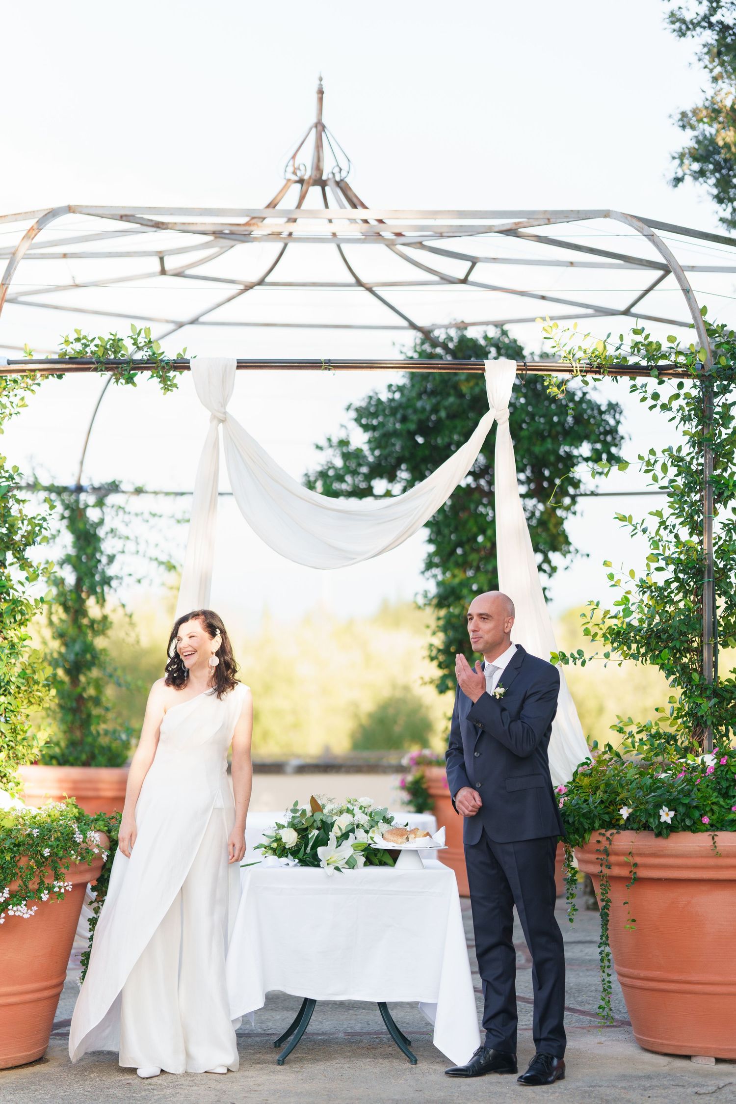 Wedding party poses under white draped canopy with terra cotta planters in outdoor garden setting.