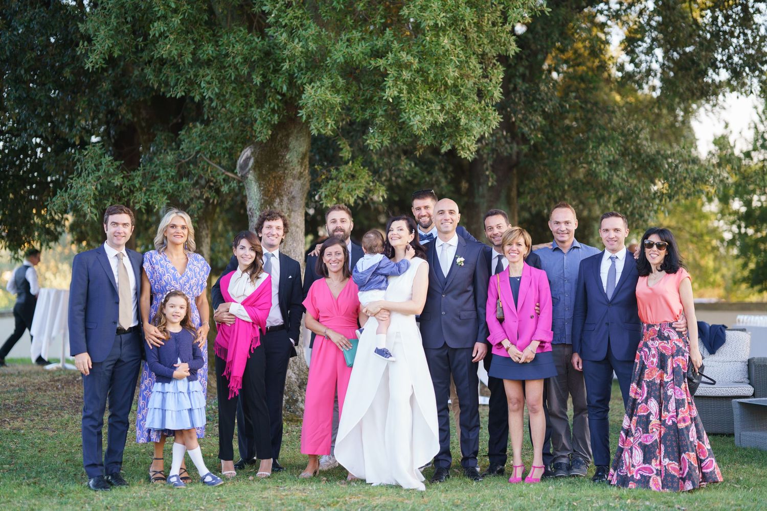 Wedding party and guests in formal attire pose together under trees for group photo.