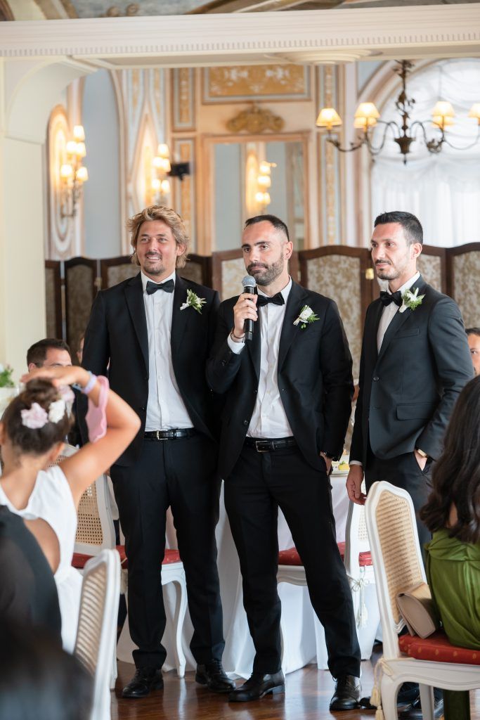Wedding party in formal black tuxedos posing against elegant ballroom backdrop.