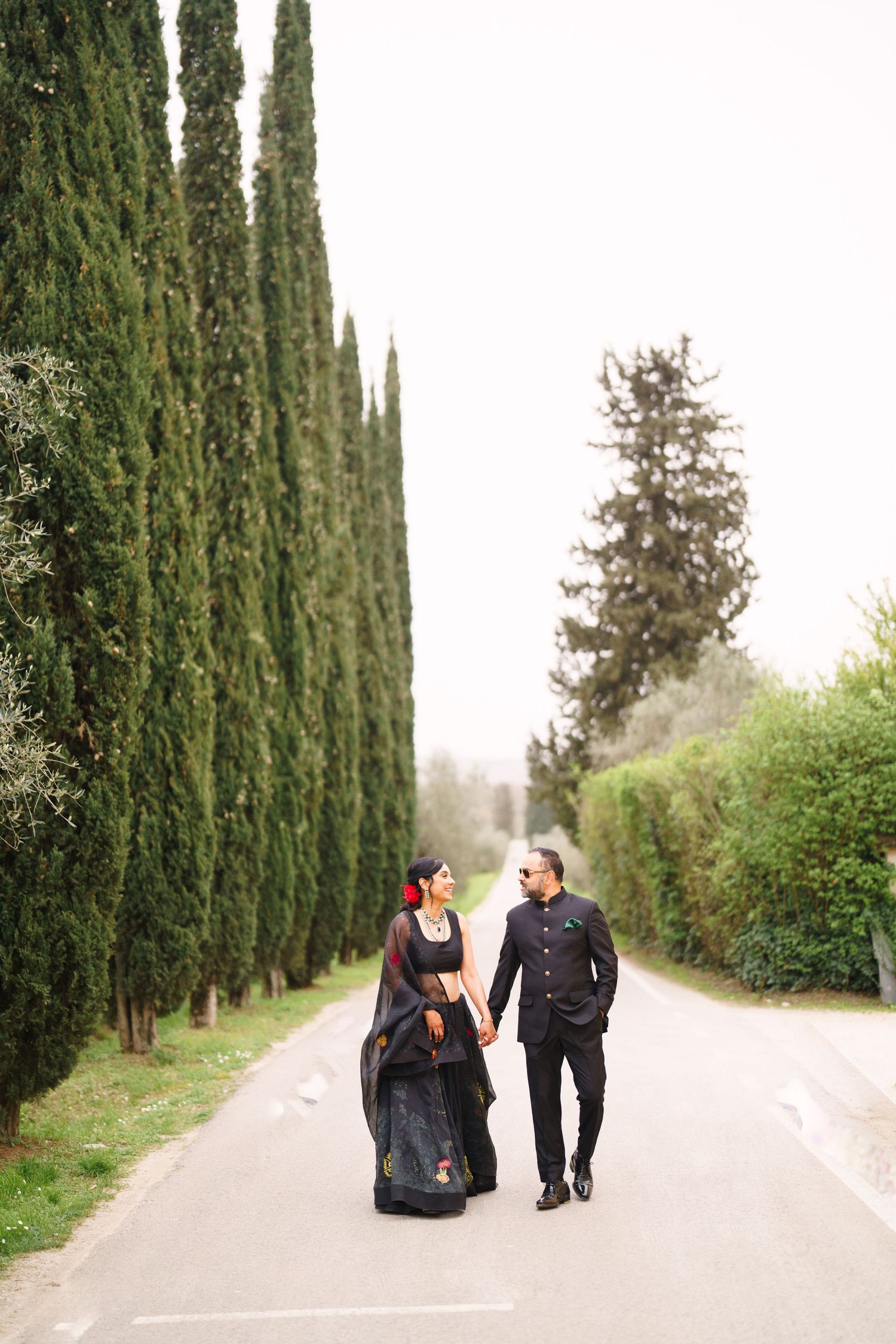 Couple walking hand in hand down cypress tree-lined path in formal wear.