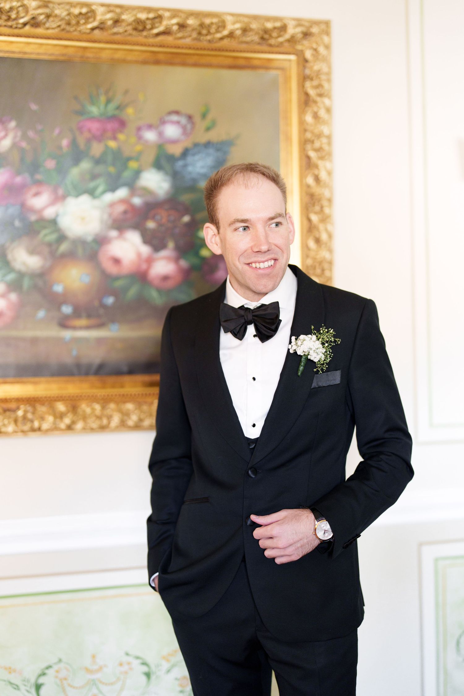 A person in a classic black tuxedo poses in front of ornate gold-framed paintings.