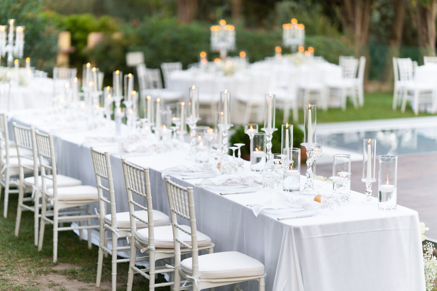 Long white dining tables set for elegant outdoor reception with silver chiavari chairs.