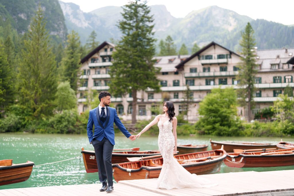 A couple poses on a dock by wooden boats with mountains and lakeside buildings in the background.