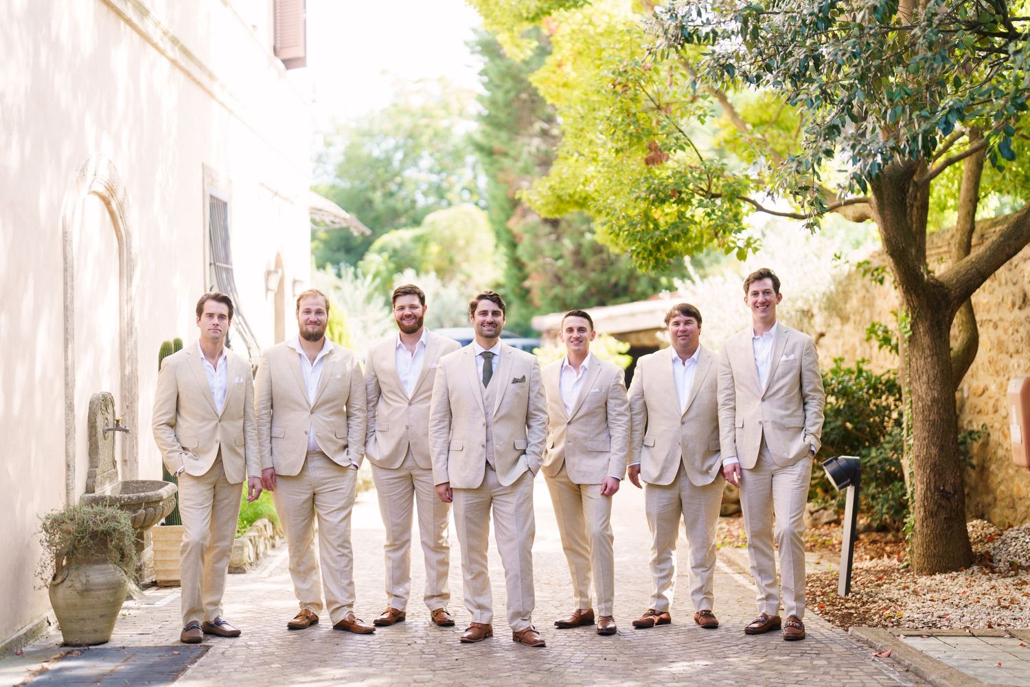 Groomsmen lineup in matching light colored suits against a garden backdrop.