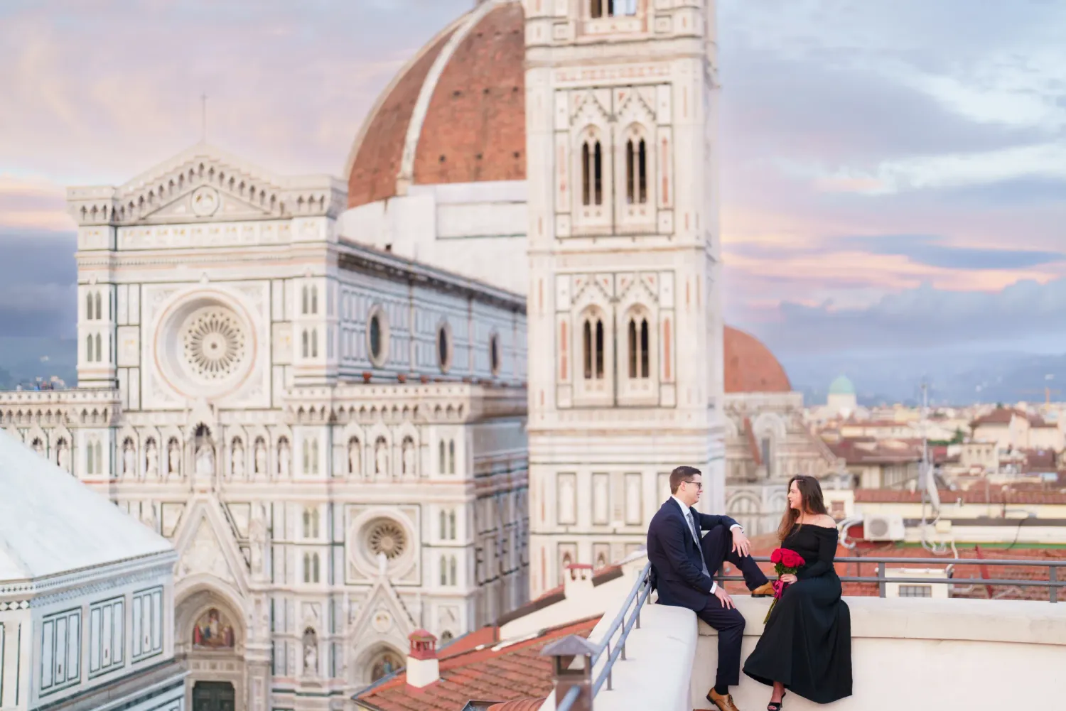 Two people sit on a rooftop overlooking the iconic Florence Cathedral and Brunelleschi's Dome during sunset in Italy.