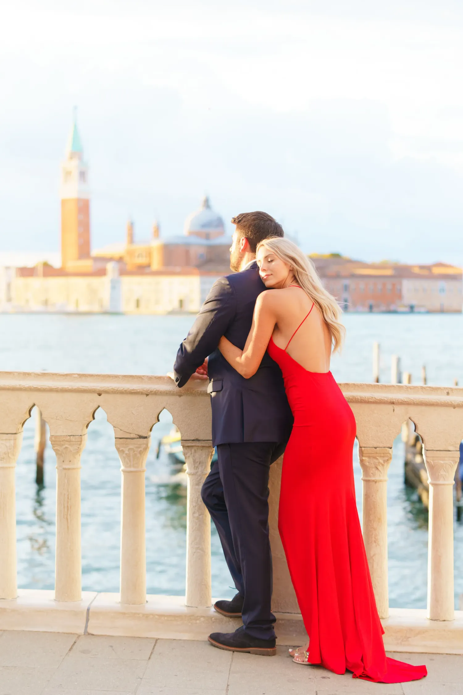A couple in formal attire embraces on a waterfront balcony overlooking Venice's iconic San Marco Basin at sunset.