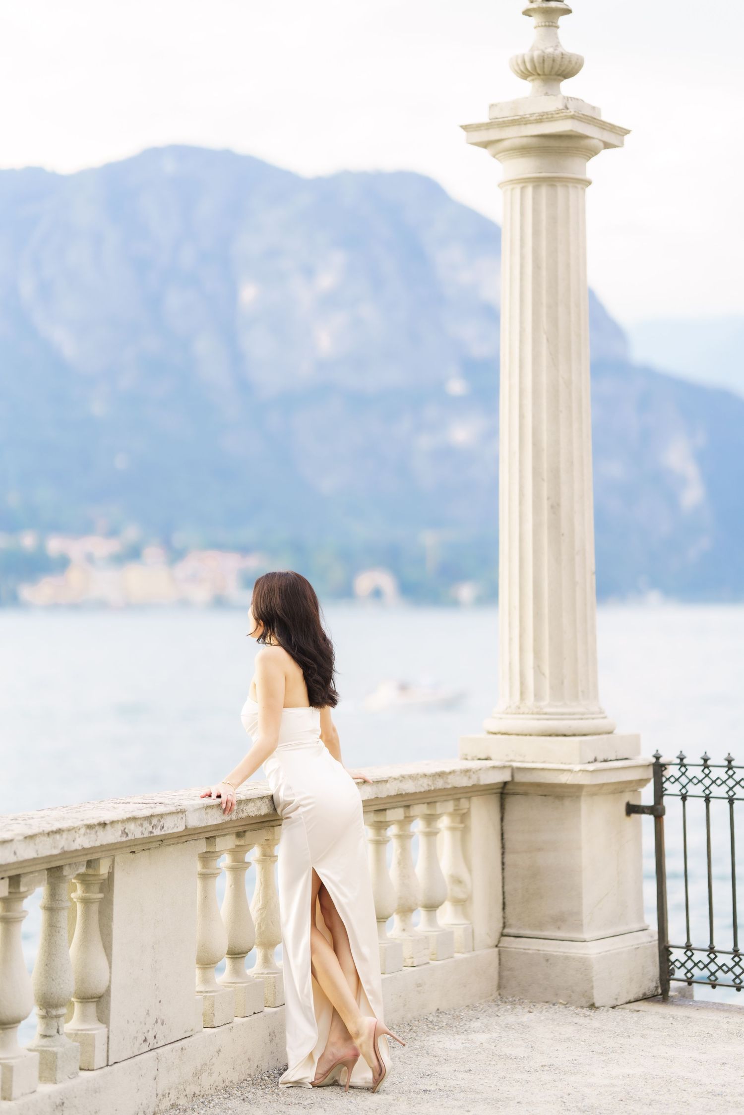 A person in a white dress stands between elegant stone pillars on a balcony overlooking a scenic mountain lake view.