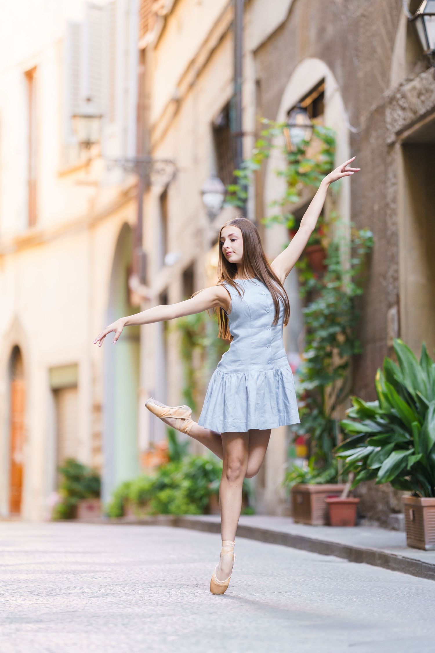 Ballet dancer performs graceful poses in a charming European alleyway with potted plants and stone architecture.