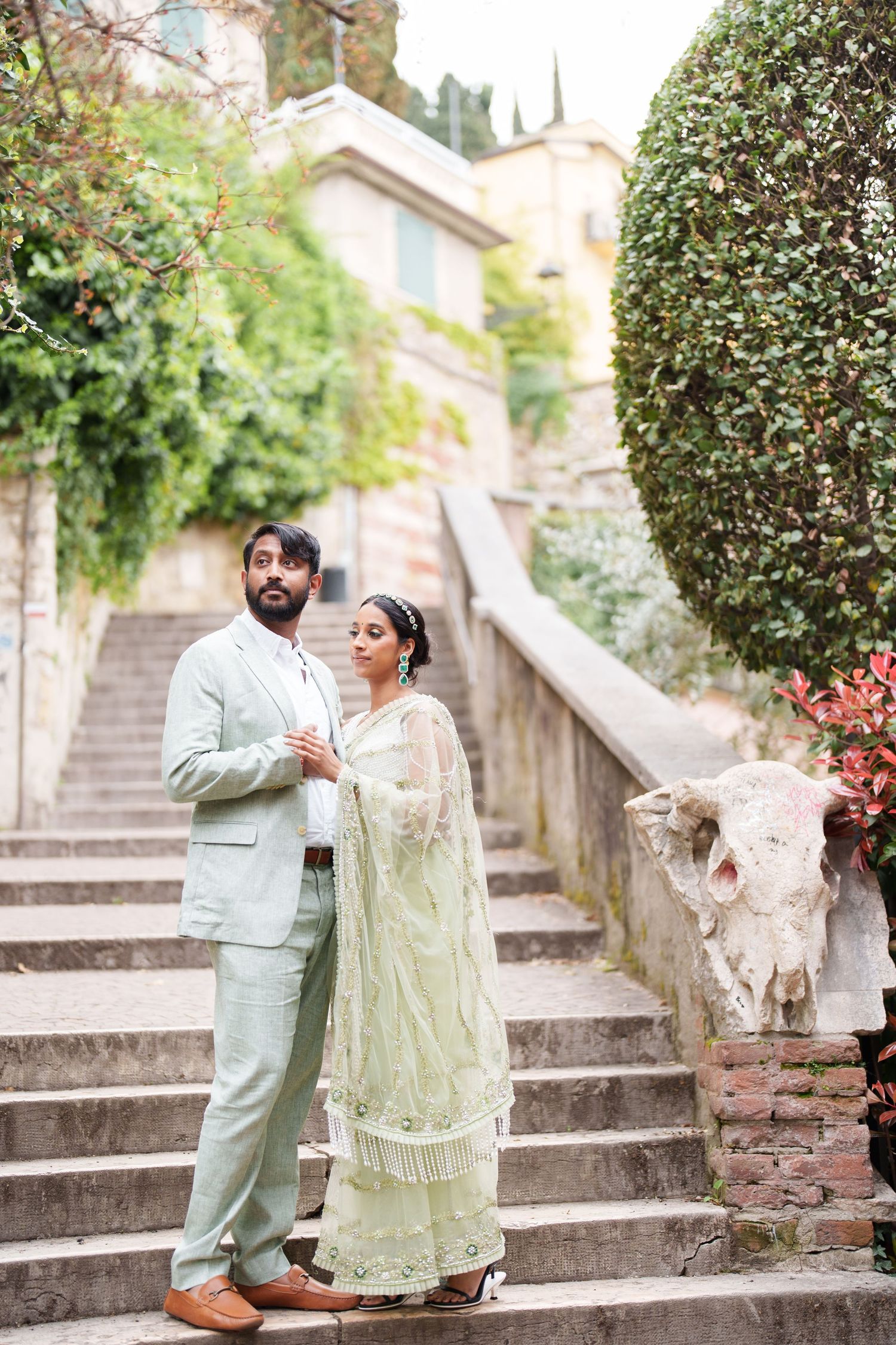 photographer verona Couple in elegant attire shares romantic moments on stone steps surrounded by greenery and blooming flowers.